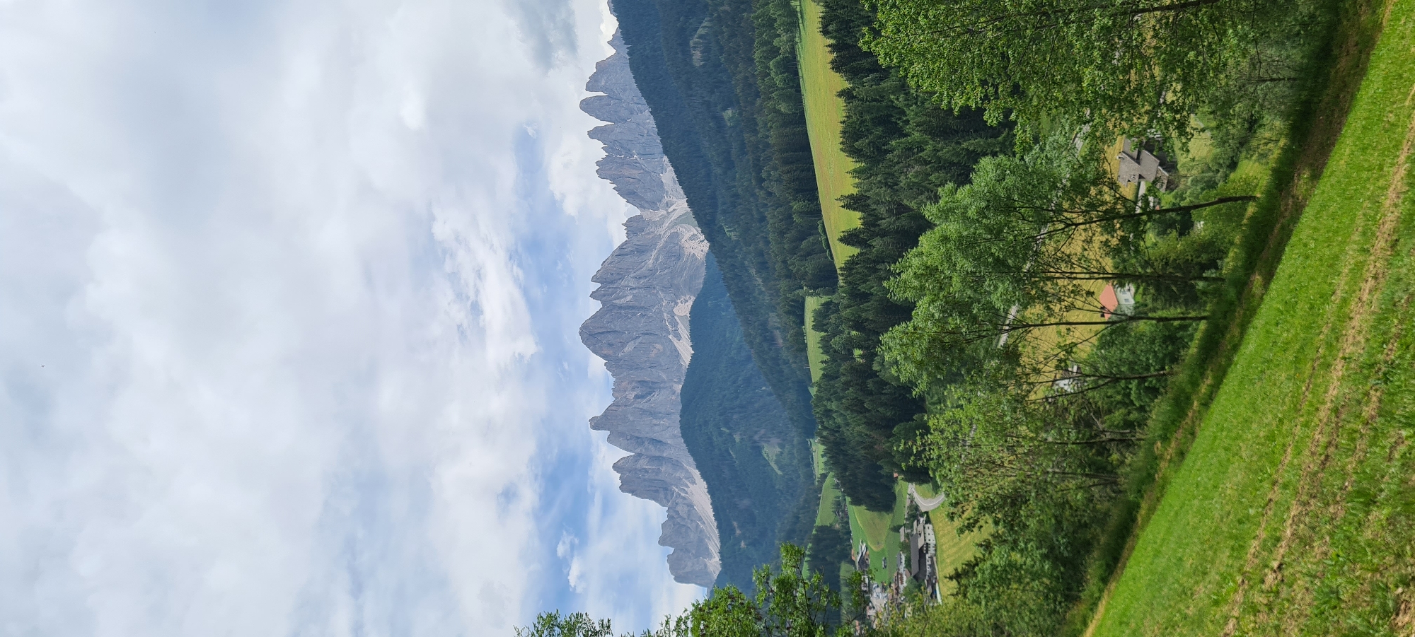 The image captures a picturesque landscape in St. Magdalena - Santa Maddalena, Italy. A jagged mountain range dominates the background, with its peaks piercing a sky filled with scattered clouds. The mountains are covered in a mix of rock faces and forested slopes. In the mid-ground, a valley unfolds, featuring a dense evergreen forest that gives way to rolling green hills and scattered houses. A winding road can be seen in the valley, connecting the small settlements. The foreground is characterized by a steep, grassy slope, with several trees dotting the landscape. The overall scene conveys a sense of natural beauty and tranquility.