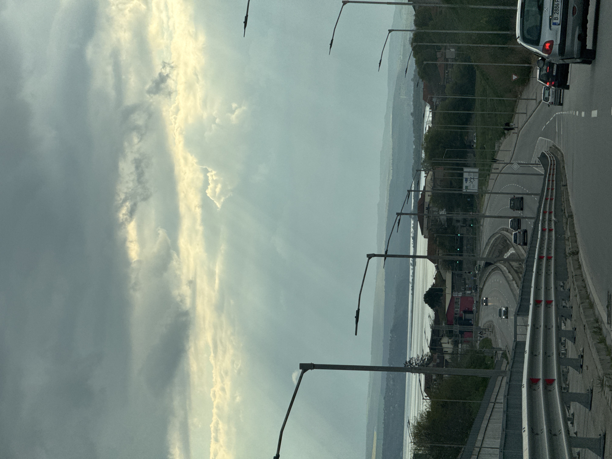 The image displays a coastal scene, likely in Varna, Bulgaria, captured from a moving vehicle. The perspective looks out over a curving road towards a vast body of water and distant land under a dramatic, partially cloudy sky.
The upper portion of the frame is dominated by the sky, characterized by a stark contrast between dark grey clouds and brilliantly lit, golden-hued clouds where sunlight penetrates, creating visible rays. This lighting suggests either early morning or late afternoon.
In the foreground and right side, a multi-lane road is prominent, equipped with metal guardrails, white lane markings, and tall streetlights. Several cars are visible on the road, indicating active traffic. In the upper right, the rear of a silver vehicle, possibly a Peugeot model, is partially seen, clearly displaying the Bulgarian license plate "B 2886 P". Other vehicles are present further along the road, some with illuminated headlights. A person can be observed walking on a sidewalk adjacent to the road in the mid-ground. Road signs, including a triangular yield sign, are also visible, though most text is illegible.
To the left of the road, a wide expanse of calm water, presumably the Black Sea, stretches into the distance. Beyond the water, hazy silhouettes of distant land or mountains complete the scenic backdrop. Along the immediate coastline and hillsides next to the road, there are scattered buildings, including one with a red roof, interspersed with lush green foliage. The scene combines natural grandeur with urban infrastructure, showcasing a dynamic interplay between sky, sea, and bustling roadway.