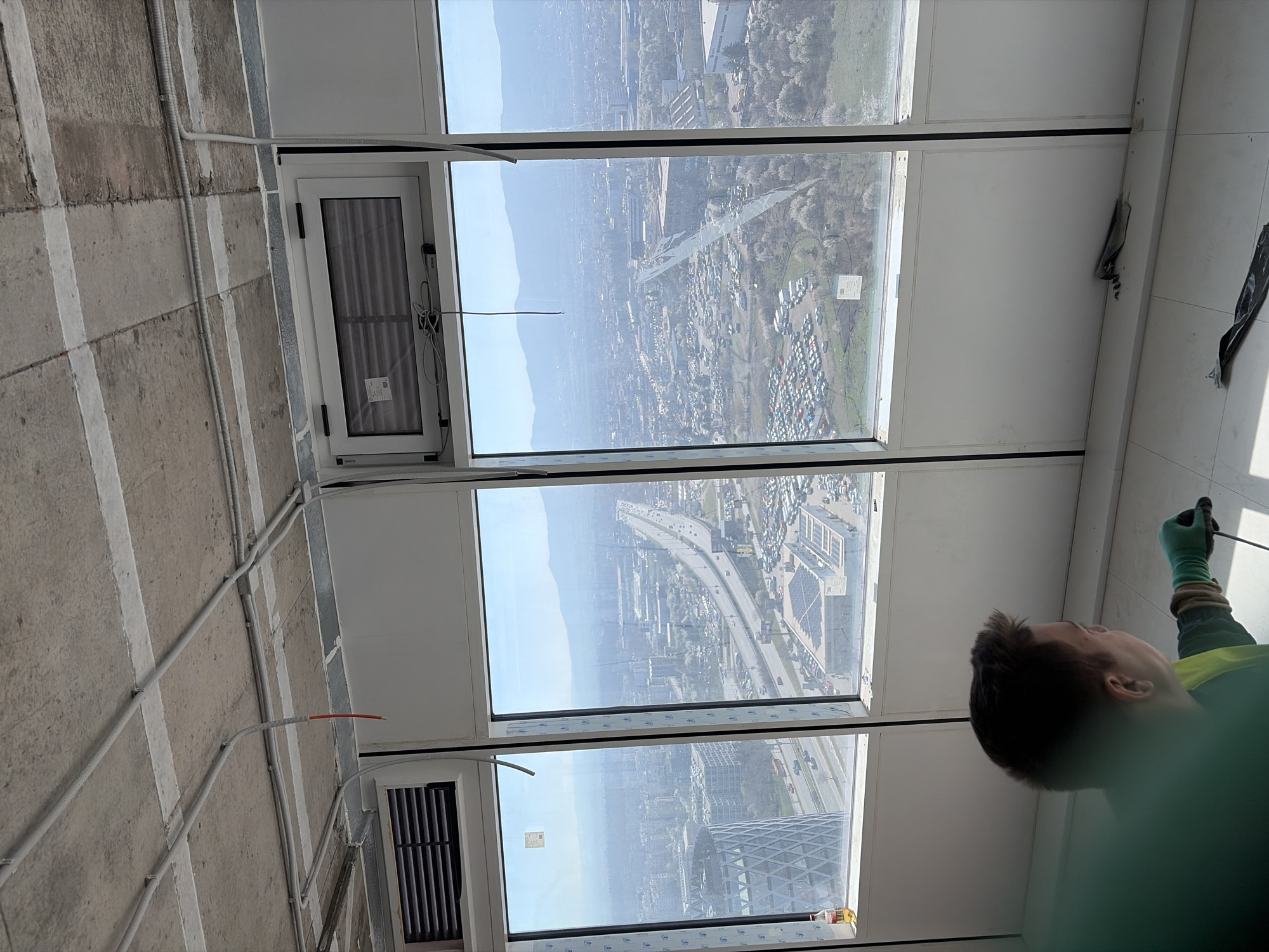 A slightly high-angle, rotated, and mostly obstructed indoor view shows a person doing work on the interior of a building in Sofia, Bulgaria, with a multi-story view to the outside. The person, partially visible at the lower right, wears a green-yellow safety vest and green gloves. They hold a thin black object, possibly a tool or cable. The foreground and right side of the image are dominated by the worker's dark hair and the vest.
The view beyond the worker features a multi-pane window revealing an expansive cityscape, presumed to be Sofia, Bulgaria. The landscape includes buildings, roadways, and patches of green space. The presence of visible highways and dense urban development confirms the location.
The window installation features both open and closed sections. Some windows have closed Venetian blinds. The building's interior shows gray and white structural elements, including conduits and wall panels. There is also a bottle of glass cleaner near the lower right corner of the bottom window, showing more glass-cleaning activity.