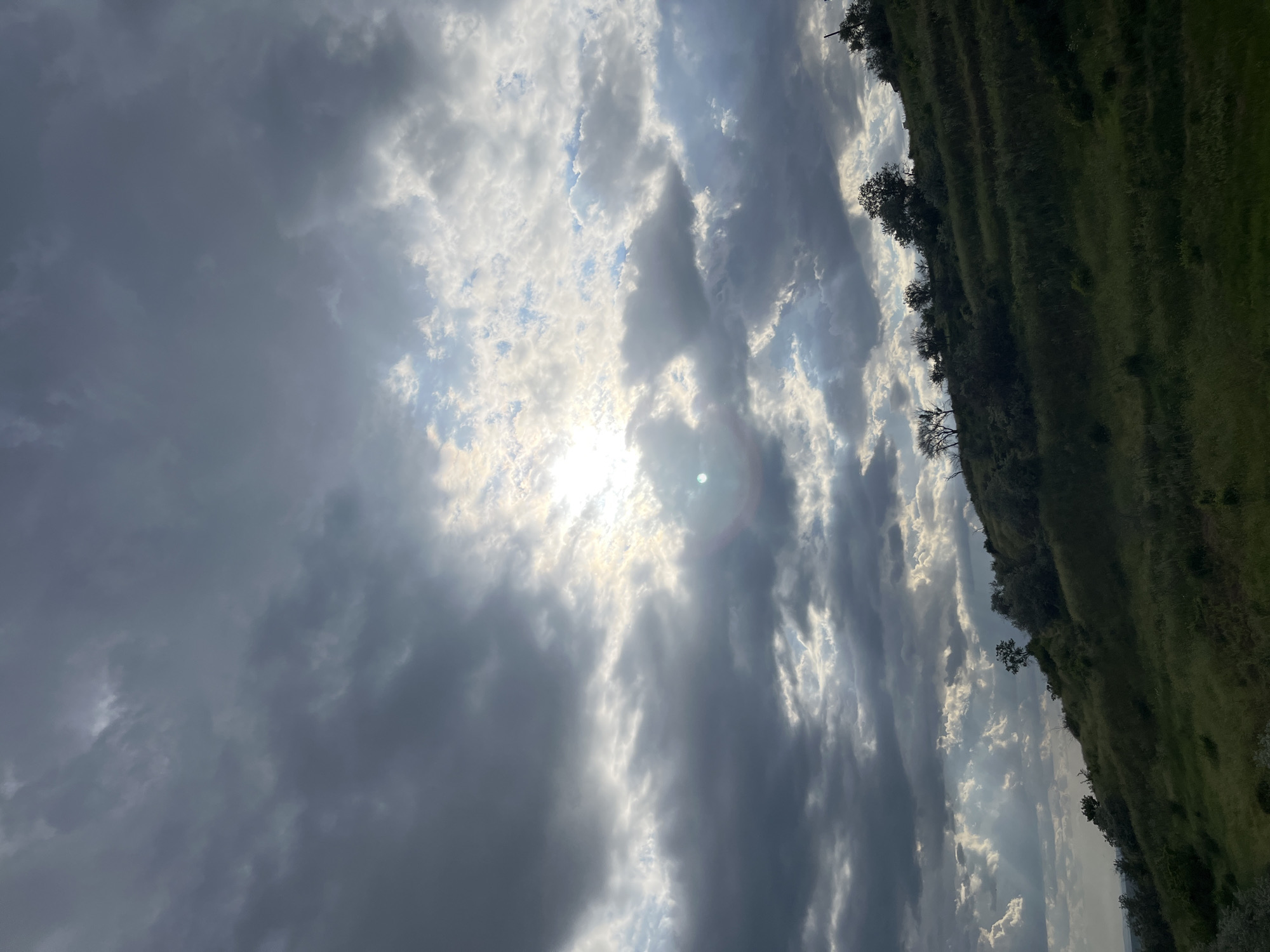 The image presents a view of the sky dominated by a mix of dark, gray storm clouds and patches of lighter, white clouds. The sun, although partially obscured, is visible through a break in the clouds, creating a bright spot. To the right, a green hillside slopes upwards, dotted with trees along its edge. The image is rotated to the side, and the location in Varna, Bulgaria, is undetermined from this single image. No people are visible.
