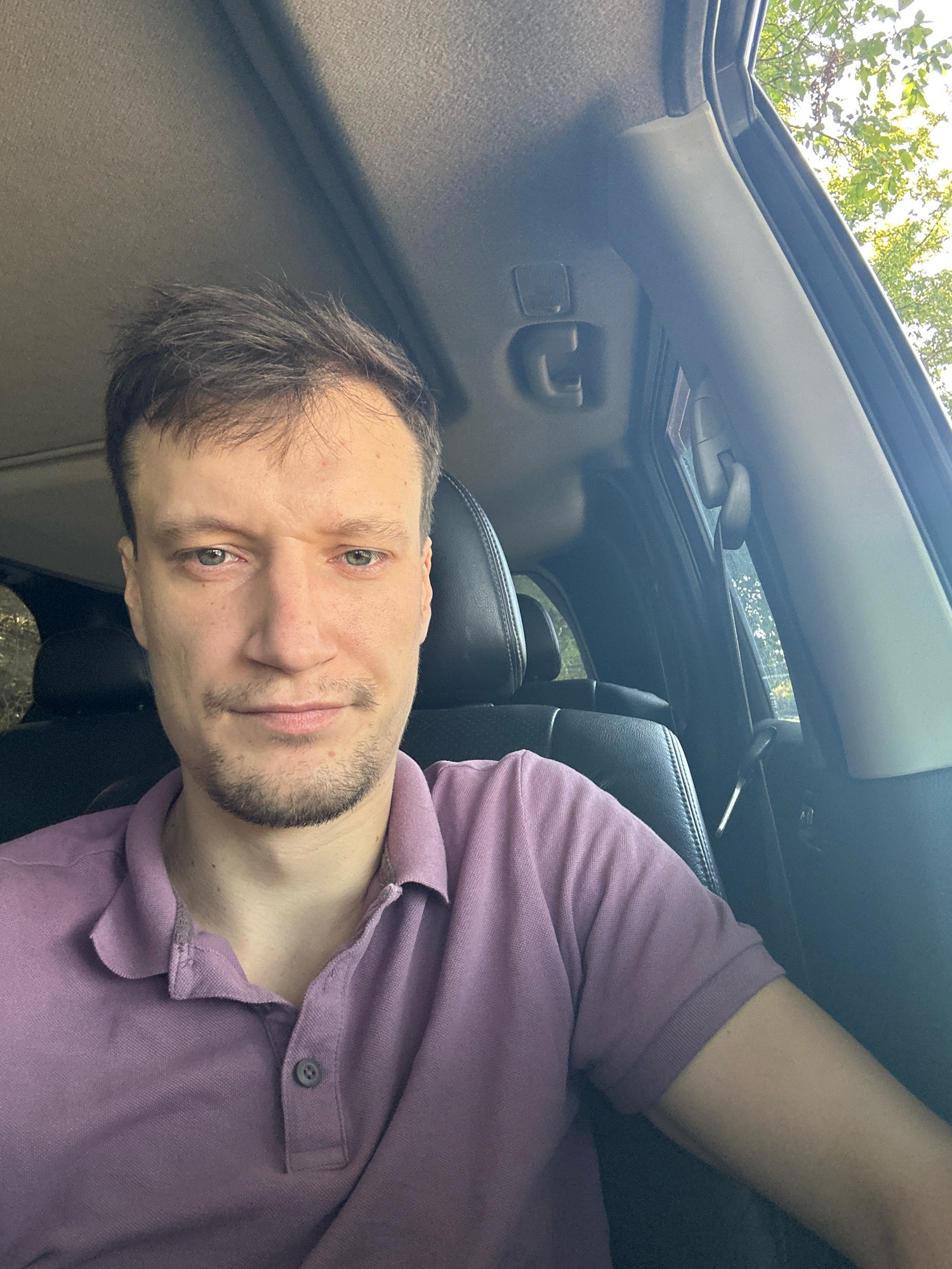 A man is sitting in the driver's seat of a car. He is wearing a purple polo shirt. The interior of the car is visible, including the dashboard, steering wheel, and passenger seat. Outside the car, trees and foliage can be seen. The photo was taken outdoors during the daytime. The scene appears to be in a rural or suburban area, possibly in or near Makiv, Ukraine, given the visible greenery. There is no indication of any activity beyond the man sitting in the vehicle.