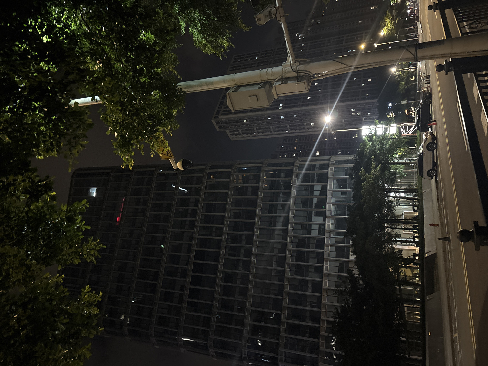This is a nighttime photograph taken in Haizhu District, China. The scene features a large, modern skyscraper with a facade of dark glass and numerous windows, reflecting the ambient city lights. In the foreground, lush green trees with dense foliage obscure parts of the view, suggesting an urban park or streetscape with significant greenery.
A prominent overhead structure, likely part of a traffic light or street lamp system, spans across the frame. It appears to be made of metal and supports electrical boxes or fixtures. Below this structure and to the right, a street is visible with parked cars, including a dark-colored sedan and a lighter-colored car. The street is illuminated by streetlights, creating streaks of light and reflections on the wet-looking pavement, hinting at recent rain or a damp environment.
The overall impression is one of a busy, developed urban environment at night, with a contrast between the man-made structures and the natural elements of the trees. The lighting is dim and atmospheric, characteristic of a city after dark. No people are clearly visible, and the focus is on the architecture, infrastructure, and the overall ambiance of the nighttime cityscape. The image is captured from a slightly upward-angled perspective, looking towards the buildings and the sky.