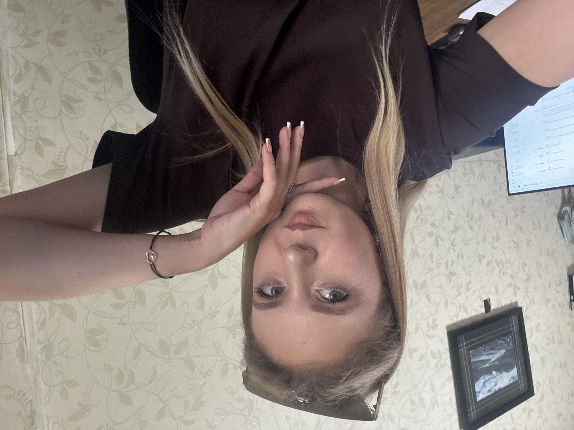 A young Caucasian woman with blonde hair pulled back and sunglasses perched on her head is captured in a selfie. She is wearing a dark brown t-shirt. Her left hand is propped on her chin, with white-tipped fingernails visible. A black bracelet with a small heart charm is on her right wrist. She is indoors, with a floral patterned wallpaper behind her. To her left, a framed picture of a landscape hangs on the wall, and a computer monitor is visible on a desk. The scene appears to be in a domestic or office setting in Unknown, Unknown.