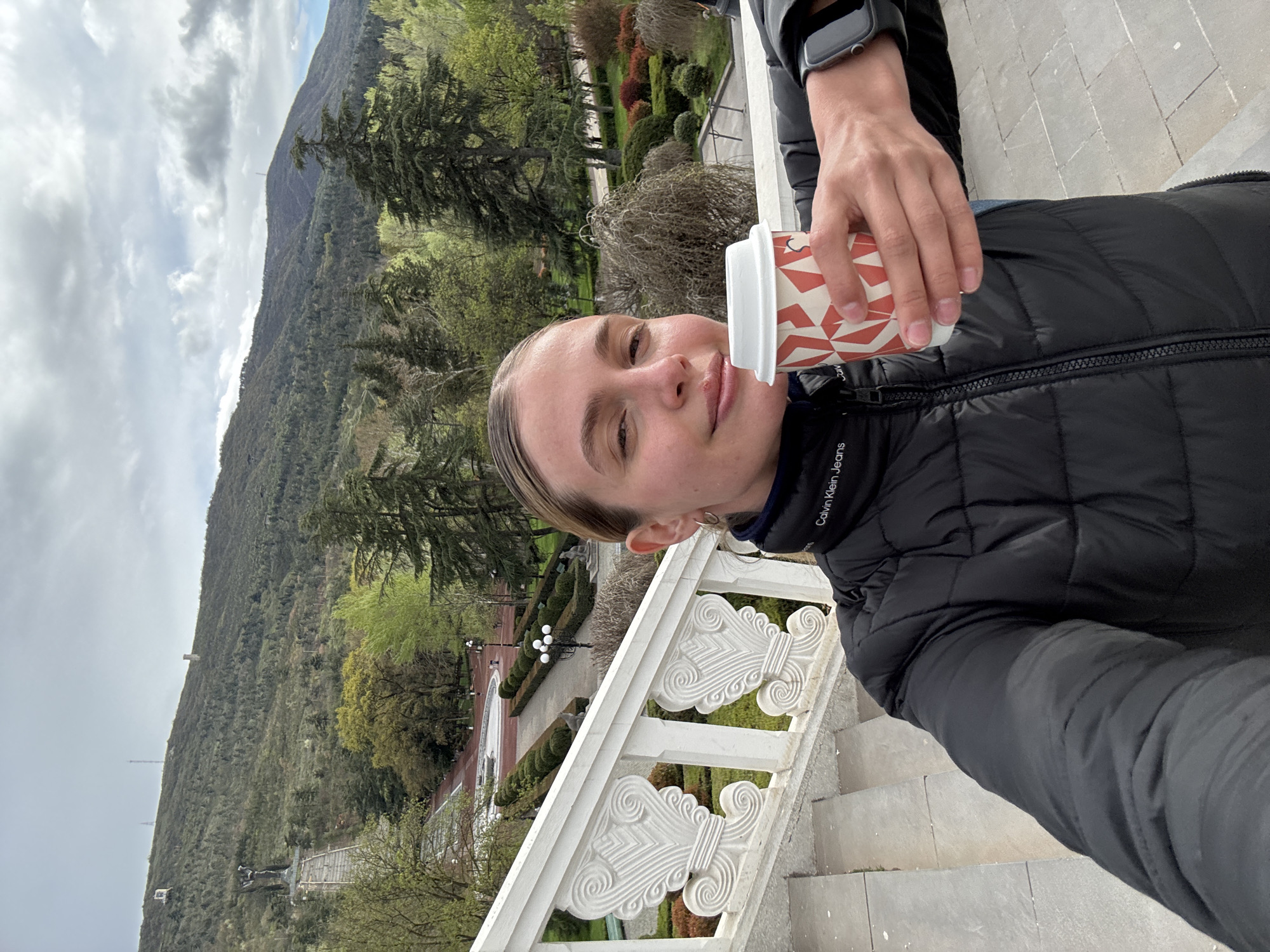A woman takes a selfie outdoors on a stone staircase with an elaborate white railing, holding a paper coffee cup with a red and white geometric pattern. She is wearing a black puffer jacket with the words "Calvin Klein Jeans" visible on the arm. Behind her and to the left is a view of a tree-covered mountain landscape under a cloudy sky. In the distance, a statue can be seen in a landscaped garden. This image was taken in Tbilisi, Georgia.