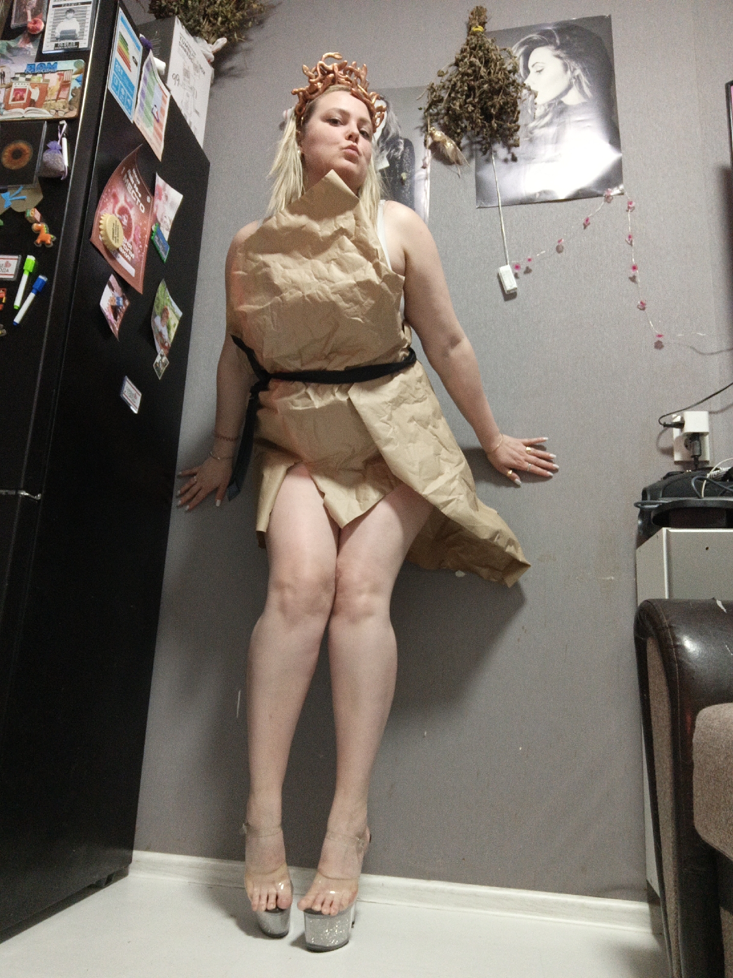 A woman with blonde hair is depicted indoors in Markovskiy, Russia, striking a pose in a creative costume. She wears a dress fashioned from crumpled brown paper bags, cinched at the waist with a black belt or sash. On her head, she sports a golden, snake-like crown, evoking the image of Medusa. Her footwear consists of clear platform high heels with glittery soles. She leans against a grey wall and a large black refrigerator, with her hands resting on the surfaces. Her expression is a slight pout.
The background features a domestic setting. The black refrigerator is covered with various magnets, photos, and notes, some of which show illegible Cyrillic text, although "МЕСТО" (MESTO - place) is visible on a yellow magnet and "ЖКХ" (ZhKKh - housing and utility services) on another paper. Above the woman, a bunch of dried foliage or herbs hangs from the wall next to a black-and-white poster depicting a woman's profile. A string of fairy lights with pink decorative elements cascades down the wall. Electrical outlets and wires are visible on the right, along with a partial view of a brown upholstered armchair. The floor is light-colored with a white baseboard. The lighting suggests an indoor setting, likely under artificial light.