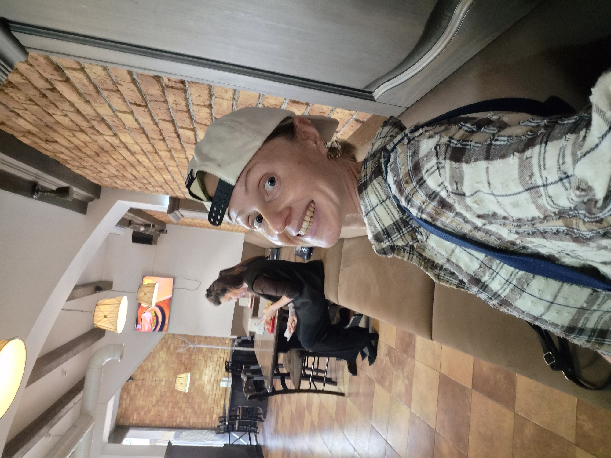 The photograph depicts a woman wearing a baseball cap and a plaid shirt. She is smiling at the camera. Behind her, another woman is seated at a table, seemingly occupied with something in front of her. The scene appears to be inside a restaurant or cafe, with tiled flooring and brick walls visible. There are hanging lights and a menu board. The photograph appears to be taken in the city of Sochi, Russia.