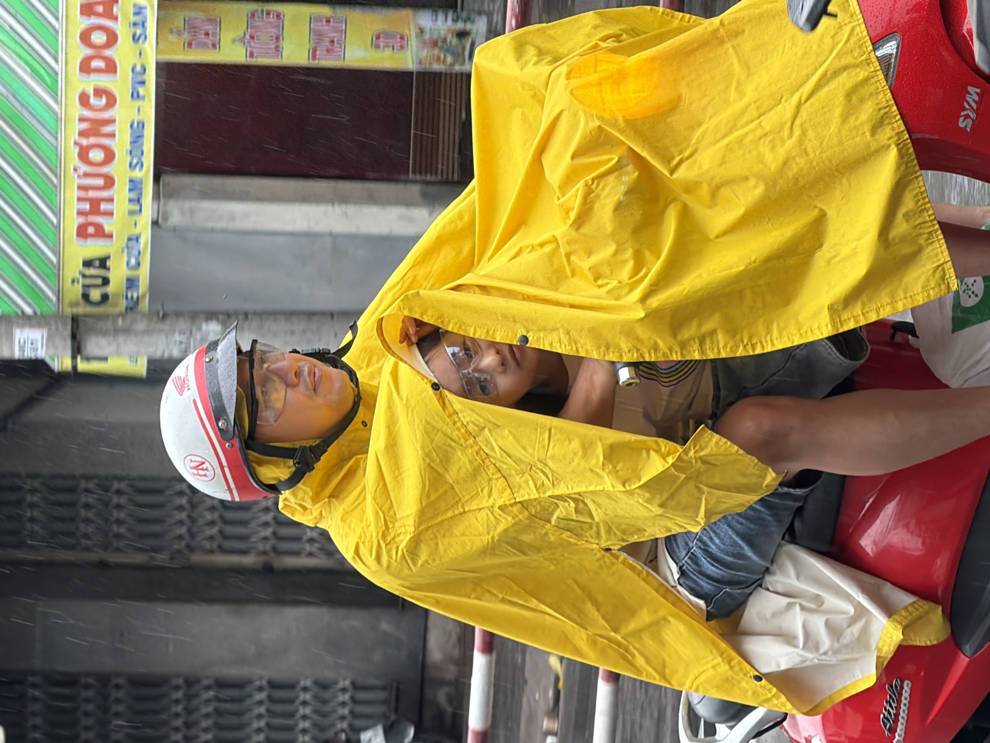 A person wearing a yellow poncho and a helmet rides a red scooter in the rain on a city street in Nha Trang, Vietnam. The person's face is obscured by the poncho, but they are wearing glasses. A child is tucked underneath the poncho with the rider. Buildings with signs are visible in the background. The street appears wet and reflects the surrounding environment.