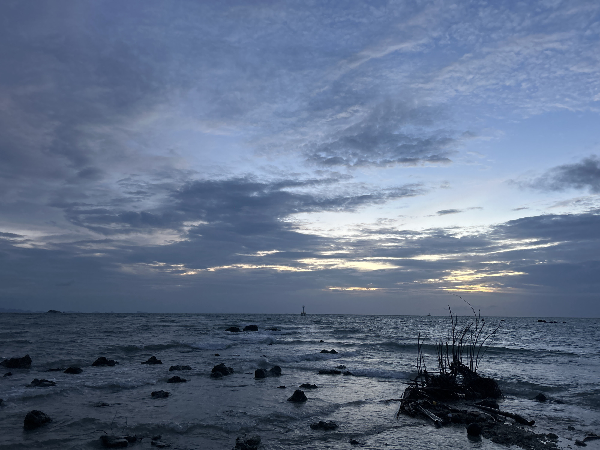 The image captures a coastal scene at dusk on Ko Pha-ngan District, Thailand. The sky is filled with a complex layer of clouds, ranging from deep blue to lighter shades with hints of the setting sun peeking through. The sea is moderately choppy, with waves breaking on a rocky shoreline. In the foreground, the water laps against dark, irregular-shaped rocks, some of which are partially submerged. A cluster of driftwood and bare branches is visible on the right side of the frame, partially in the water. In the distance, a small lighthouse or marker can be seen standing in the sea. The overall atmosphere is serene and a little moody, typical of twilight over the ocean.