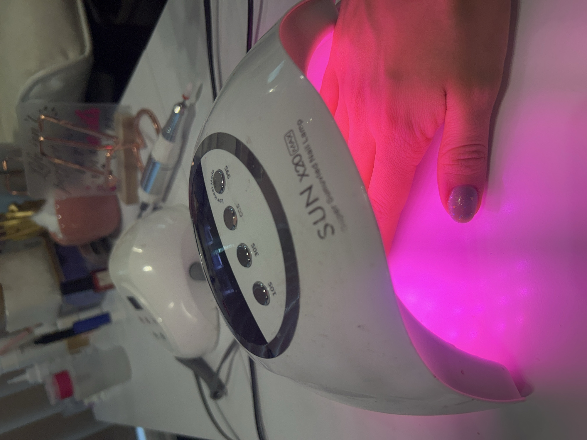 A right hand with a purple fingernail is in a white, illuminated, SUN X20 MAX nail lamp. The nail lamp has several buttons, one marked "99S Low Heat Mode." The lamp sits on a white surface with other manicure tools and supplies. The setting appears to be indoors and is possibly a nail salon. The location is indiscernible in Sofia, Bulgaria.