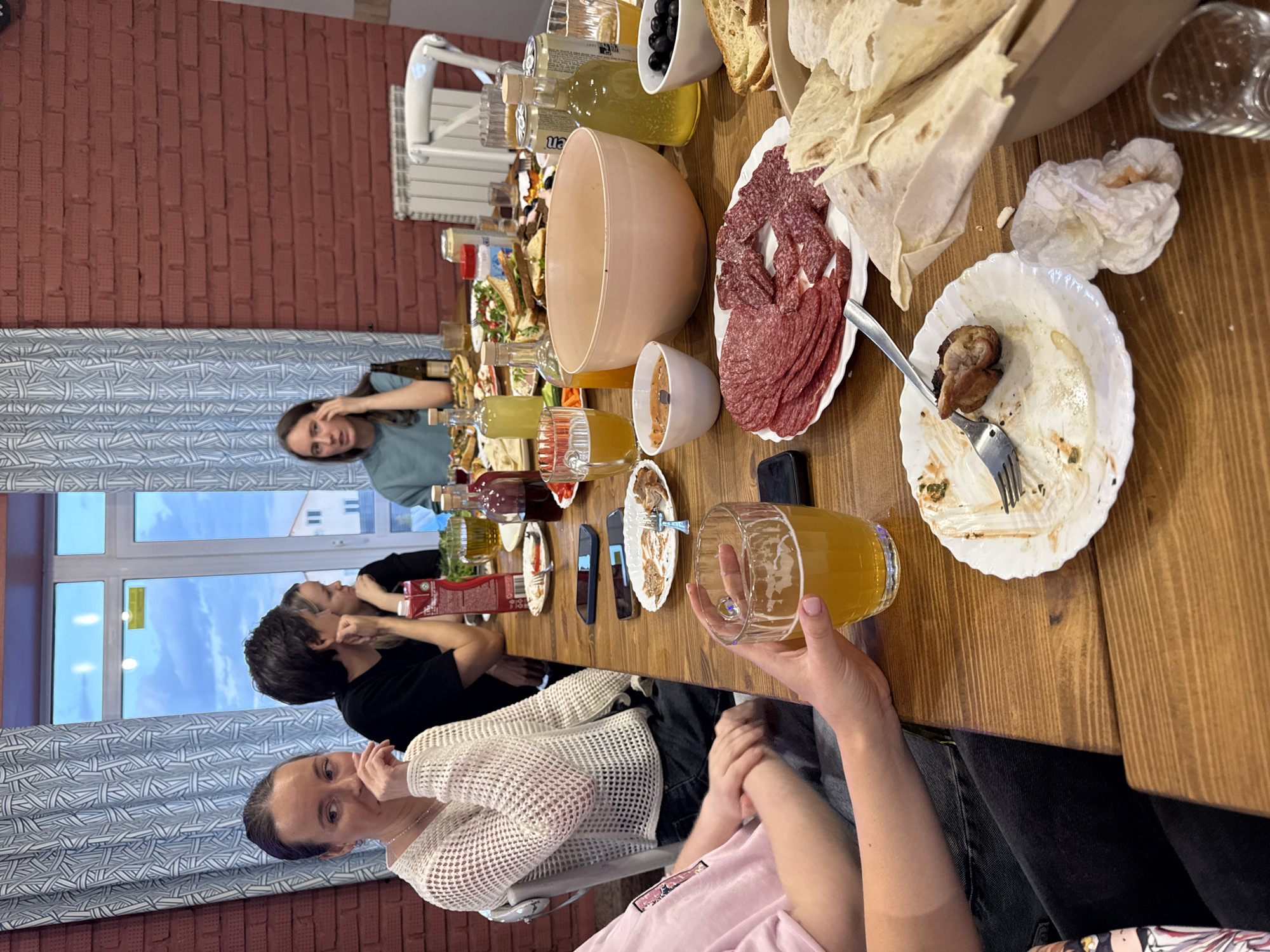 The image shows a group of people gathered around a table laden with food and drinks, likely at a gathering in Бугачёво, Russia. Several individuals are visible, including a woman in a white sweater touching her face, another woman in a pink shirt holding a glass of beer, and two individuals in black shirts near the window. On the table, there are various dishes, including plates with salami, flatbreads, olives, and leftovers, along with bottles of liquids and a large beige bowl. Some people are interacting and appear to be talking and eating. The environment includes a red brick wall and a window with a view of the sky.