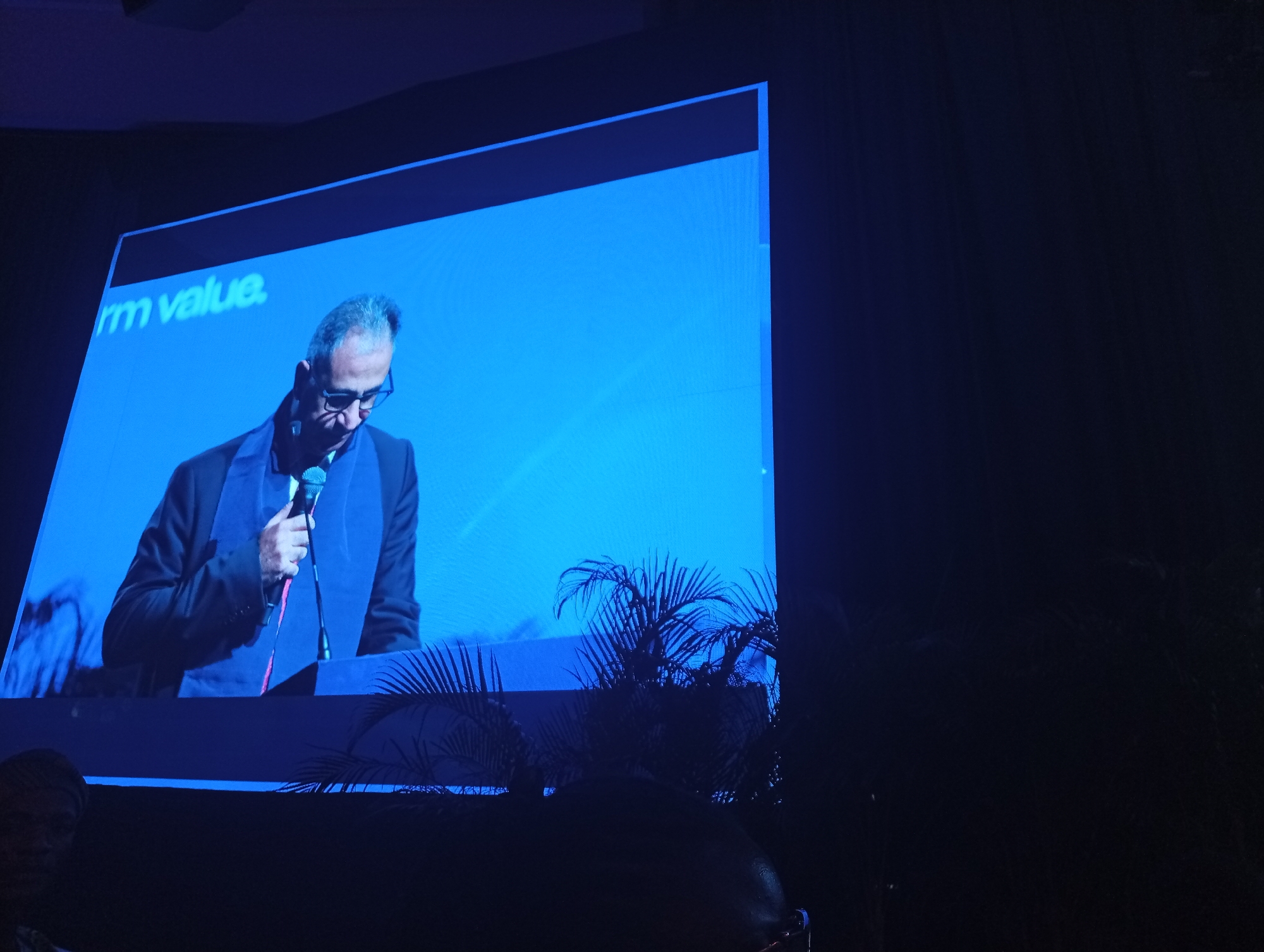 A projected image displays a man with short, gray hair, wearing glasses and a dark suit. He is holding a microphone and looking down, as if reading from a podium. The background of the projected image is solid blue and includes the text "rm value". The projector appears to be set up in front of decorative plants that are in front of a dark audience. The venue is otherwise not visible and there is no indication of a location in Itirin, Nigeria.