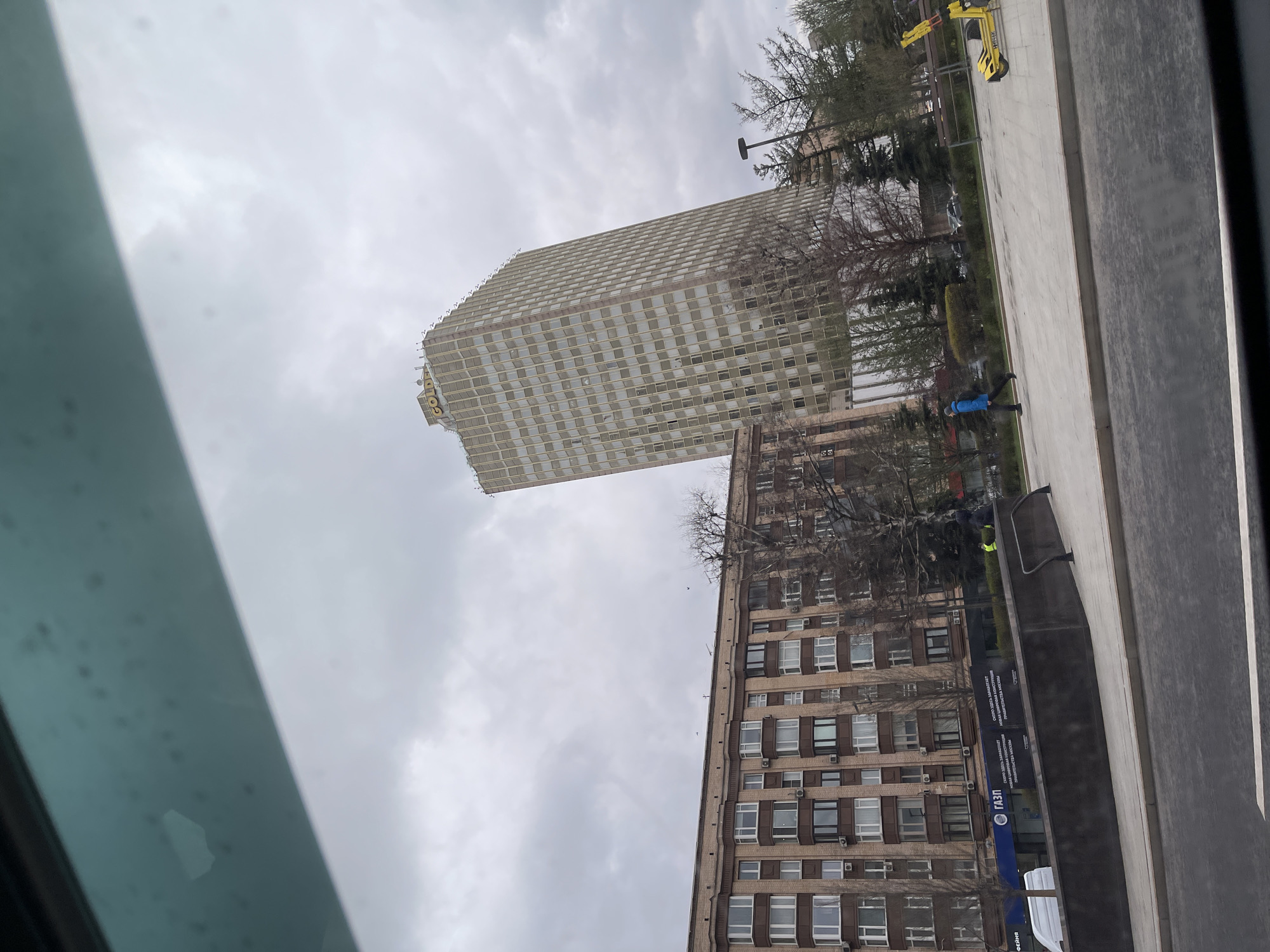 The image shows two buildings in Moscow, Russia, on an overcast day. The building on the left is a tall, rectangular, grid-patterned high-rise with the "GOLDEN" logo visible on top. To its right is a shorter, brown brick building with numerous windows and some visible air conditioning units. In front of the buildings is a grey pavement area with some people walking and a yellow scooter. A black sign with blue accents and Cyrillic script, featuring the "GAZ" logo, is present. The view is likely taken from inside a vehicle, with part of the vehicle's interior visible on the left edge of the image.