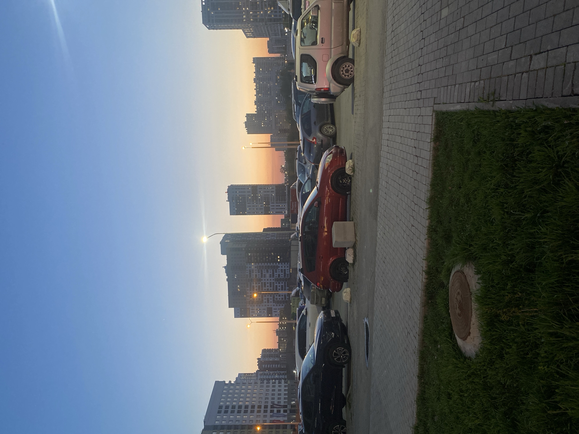 The image depicts a row of parked cars along a sidewalk with a patch of green grass to the right. A sewer access cover is set in the grass. In the background, tall buildings, typical of an urban area, rise against a sky transitioning from blue to orange at the horizon, suggesting either sunrise or sunset. Streetlights are visible between the buildings. The location is identified as a city of Yekaterinburg, Russia.