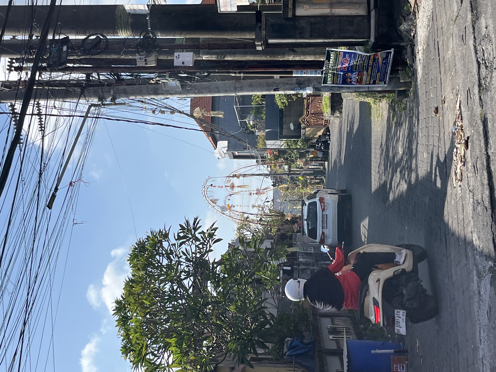 A street in Denpasar, Indonesia, is depicted on a bright, sunny day, with a clear blue sky and scattered clouds. The foreground features a person riding a beige scooter with a white helmet, black jacket, and red shirt. The scooter has a license plate that reads "DK 9281 AEK." Behind the scooter, a white SUV is visible with its license plate reading "DK 1036 ACG." The street is paved with cobblestones and lined with buildings, trees, and numerous electrical poles and wires, characteristic of Denpasar's infrastructure. Arching decorative structures, likely for a cultural event, span across the street in the mid-ground. A poster for "SMP BINTANG PERSADA" is affixed to a pole on the right side of the image. The overall scene is a snapshot of daily life in a bustling Indonesian city, with a focus on the street and its surrounding elements.