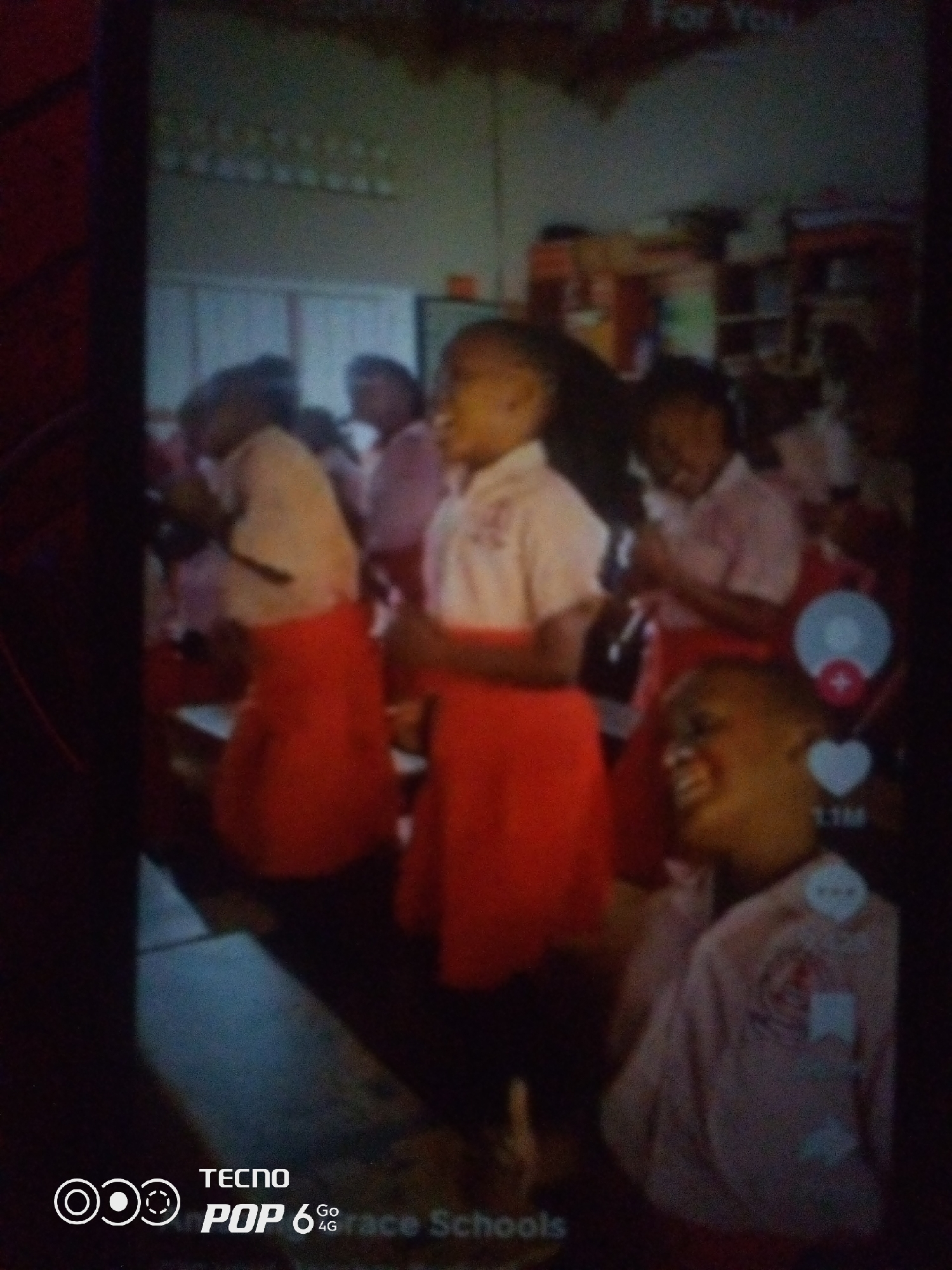 A group of young students, likely in a classroom setting, are captured in a low-resolution video still. The students are wearing similar uniforms, consisting of white tops and bright orange skirts or shorts. The room appears to be a school environment, with shelves and what might be teaching materials visible in the background. The lighting is dim, and there are overlaid icons and text from a video platform, including a heart icon with a number, indicating likes or views. The overall scene suggests a candid moment, possibly during an activity or lesson. The image's quality and the presence of video interface elements suggest it was taken with a mobile device.