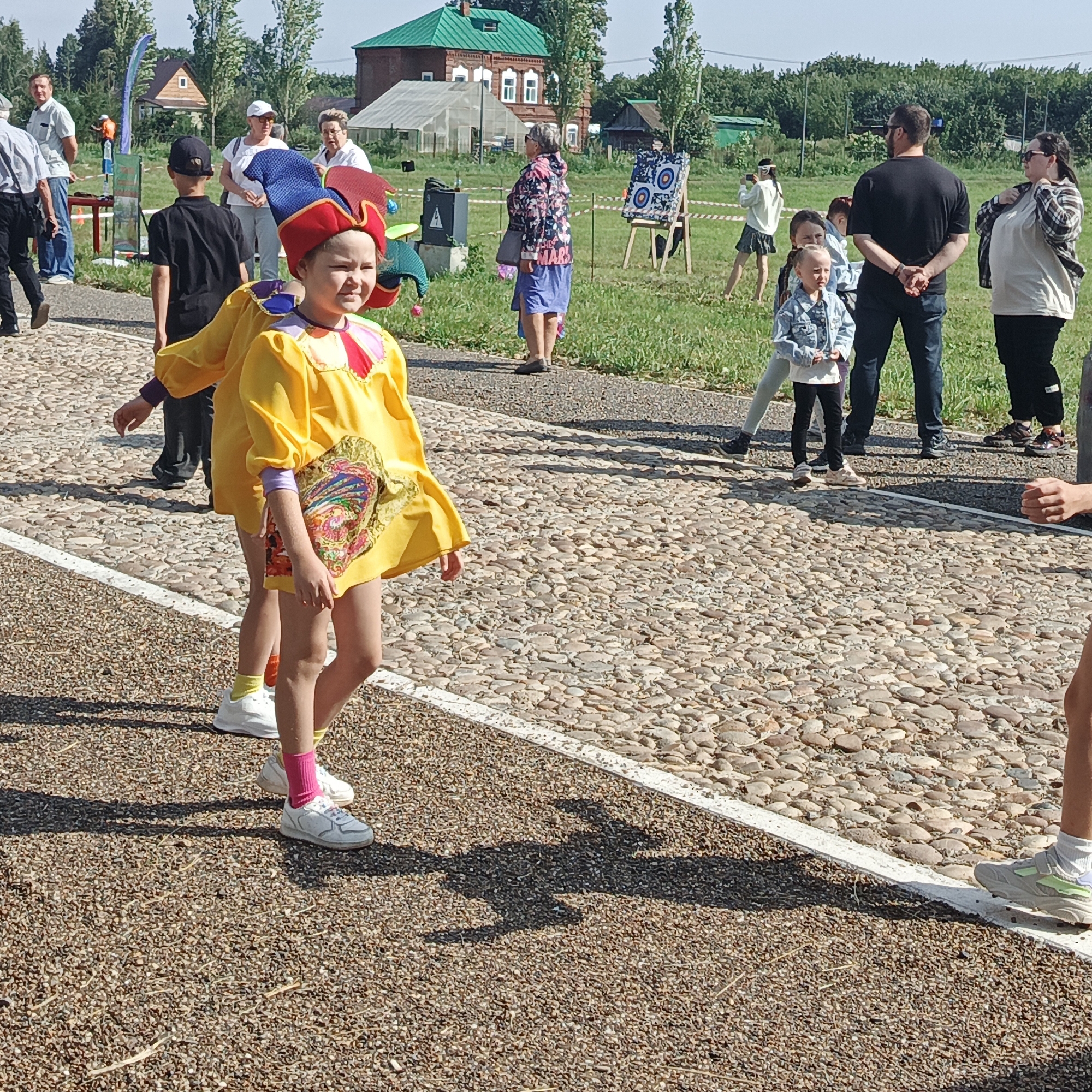 The image depicts an outdoor event with several people present on a cobblestone path. In the foreground, a child in a yellow jester-like costume with a red and blue hat is walking towards the right. Behind the child, another person in a similar yellow costume is visible. In the middle ground, a boy in a black t-shirt and cap is walking away from the camera, and further back, a group of adults and children are gathered. There is a target board with archery rings set up on the grass. In the background, there are buildings, trees, and a clear sky. The setting appears to be a park or a public space in Nikolo-Beryozovka, Russia, likely during a festive occasion or event.