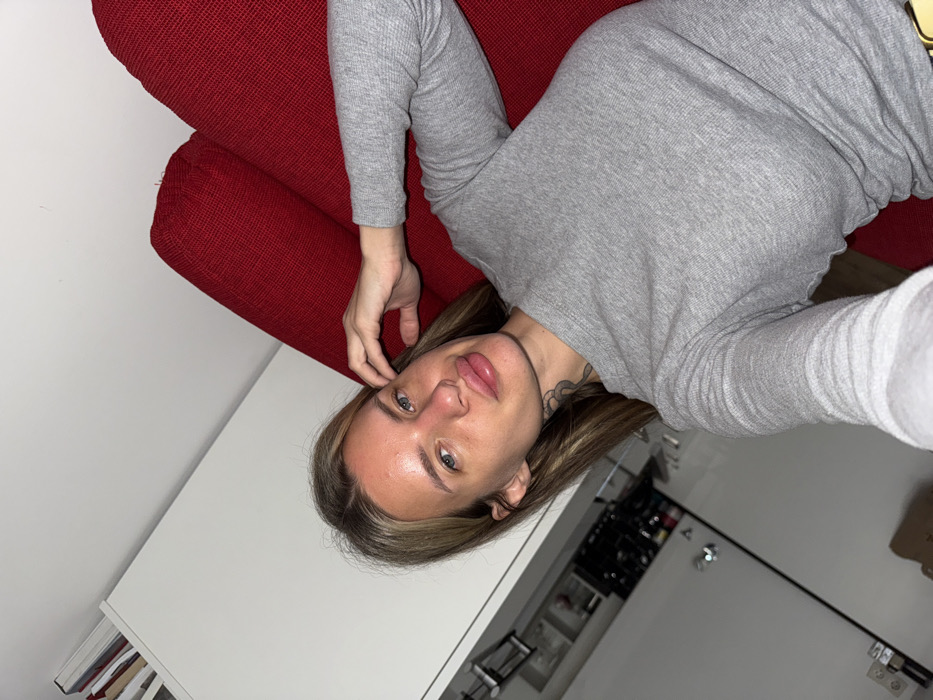 The image depicts an indoor scene, likely within a residential apartment in Berlin, Germany. A single adult female is prominently featured in a close-up, angled shot, directly facing the camera, consistent with a selfie. She possesses shoulder-length, light brown hair, blue eyes, and exhibits visibly augmented lips. A dark tattoo is partially discernible on the left side of her neck. She is clad in a form-fitting, ribbed grey long-sleeved top. Her left hand is raised, with fingers positioned near her left temple. The background to the subject's right consists of a textured red upholstered couch. To her left, a white modular shelving unit or cabinet is visible, containing multiple indistinct items such as books or decorative objects. Below this shelving, a white wall features a white double electrical outlet. The floor in the lower left foreground appears to be light-colored wood.