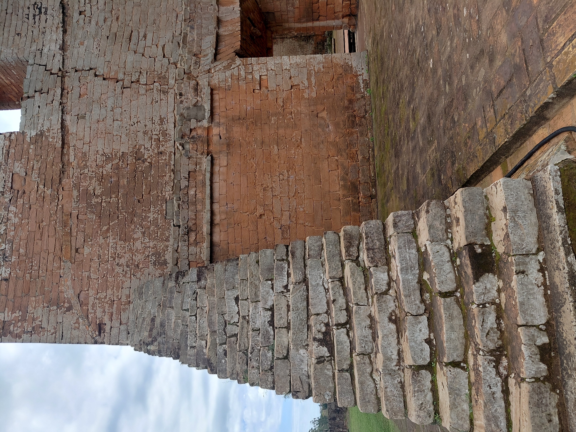 The image captures a close-up view of ancient stone steps leading up to a weathered brick structure. The steps, made of large, rough-hewn grey stones, show signs of age and wear, with moss and small plants growing in the crevices. Behind the steps, a large section of a brick wall is visible, constructed from reddish-brown bricks. This wall exhibits discoloration and erosion, suggesting it is part of historical ruins. To the right of the brick wall, an arched doorway or opening is visible, hinting at an interior space. The ground in front of the structure is paved with worn, rectangular bricks. In the background, a patch of green grass and trees can be seen under a cloudy sky. The scene is likely part of the Trinidad Jesuit Mission, a UNESCO World Heritage site located in Trinidad, Paraguay, known for its well-preserved ruins of Jesuit missions from the colonial era. There are no people visible in the image.
