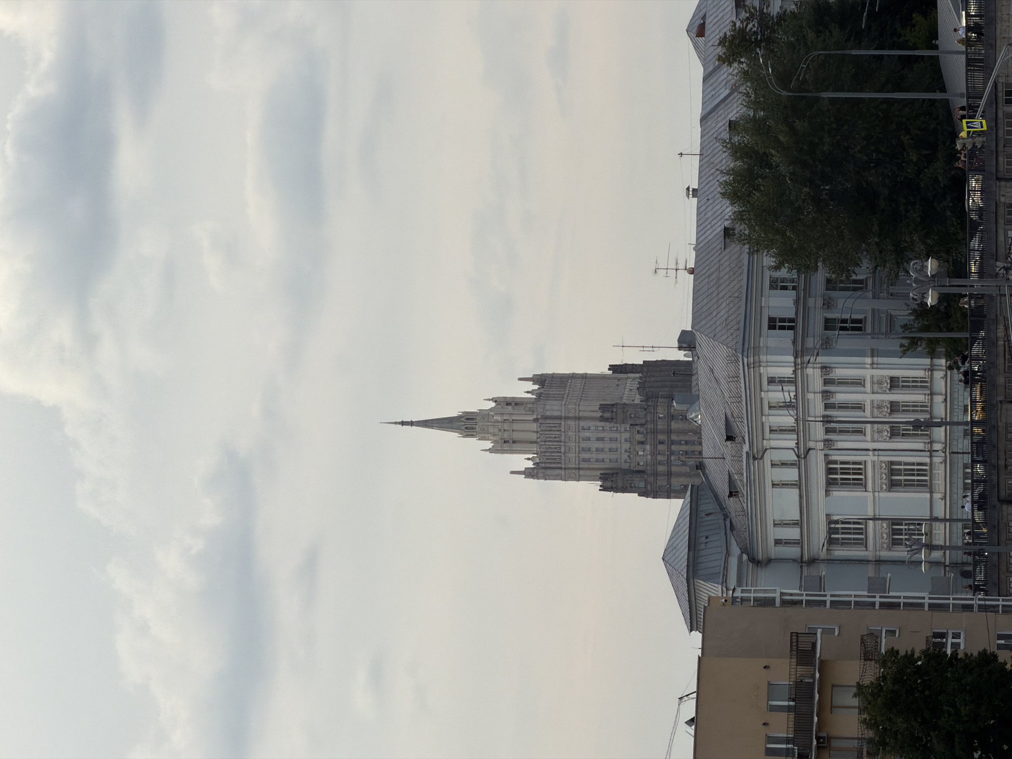 The image shows a view of Moscow, Russia, with several buildings and a cloudy sky. Dominating the background is the Stalinist skyscraper known as the Ministry of Foreign Affairs building, with its distinctive spire. In the foreground, a cluster of lower buildings is visible, including a pale yellow apartment building on the left with balconies, a light blue building with ornate white trim, and dark green trees. A bridge spans the lower part of the image, with streetlights and several people standing on it. Cars are visible on the road below the bridge. The overall atmosphere is overcast and urban.