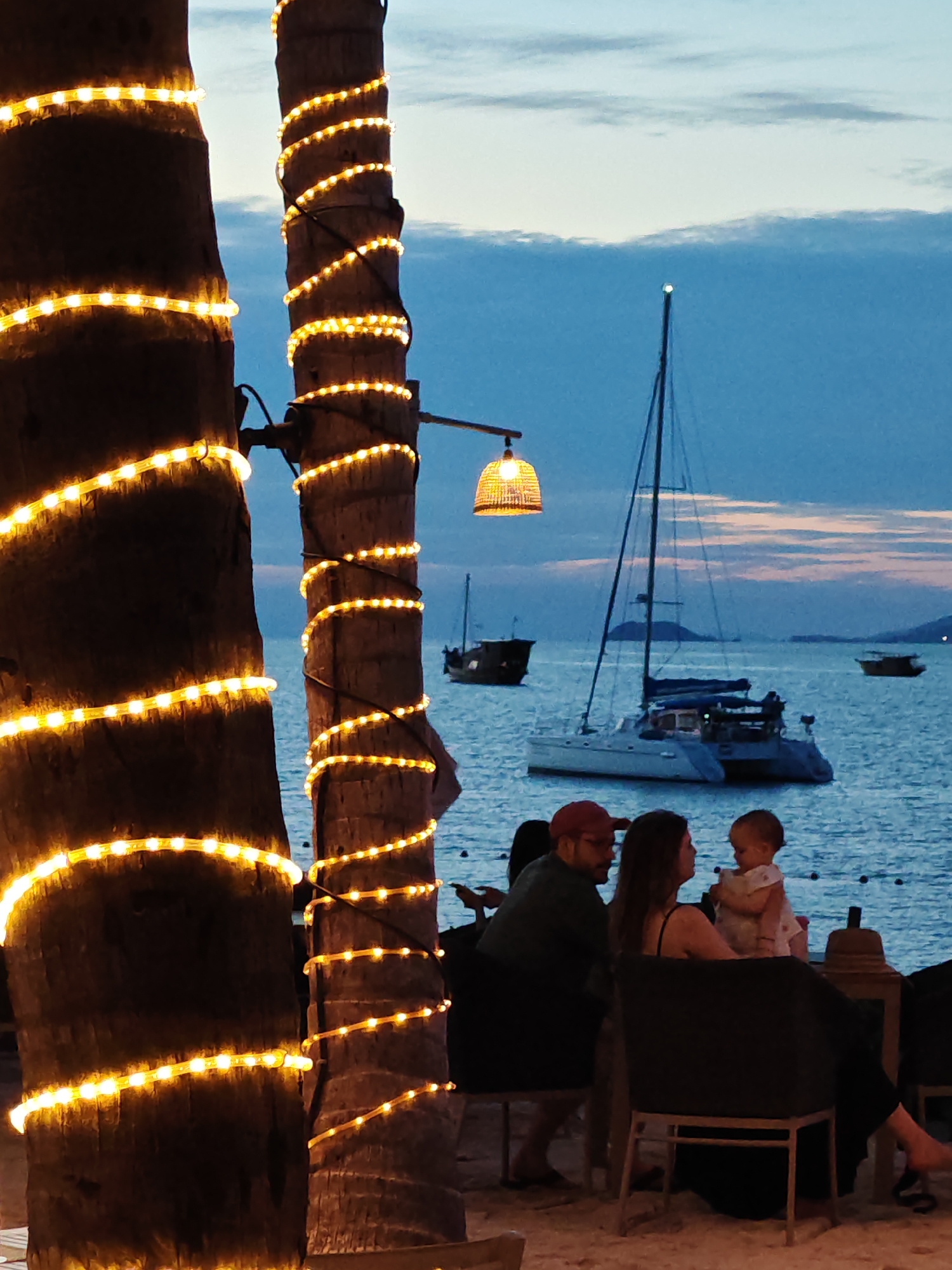 In Baan Bo Phut, Thailand, a family is enjoying a meal on a beach at dusk. Two palm trees wrapped in yellow string lights stand in the foreground, framing the scene. In the middle ground, a couple sits at a table, facing each other and looking at a baby held by the woman. The baby is looking up at the woman. The sea is in the background with several boats, including a white catamaran, sailing on the water. The sky is a dusky blue with wispy clouds. The atmosphere is relaxed and romantic.