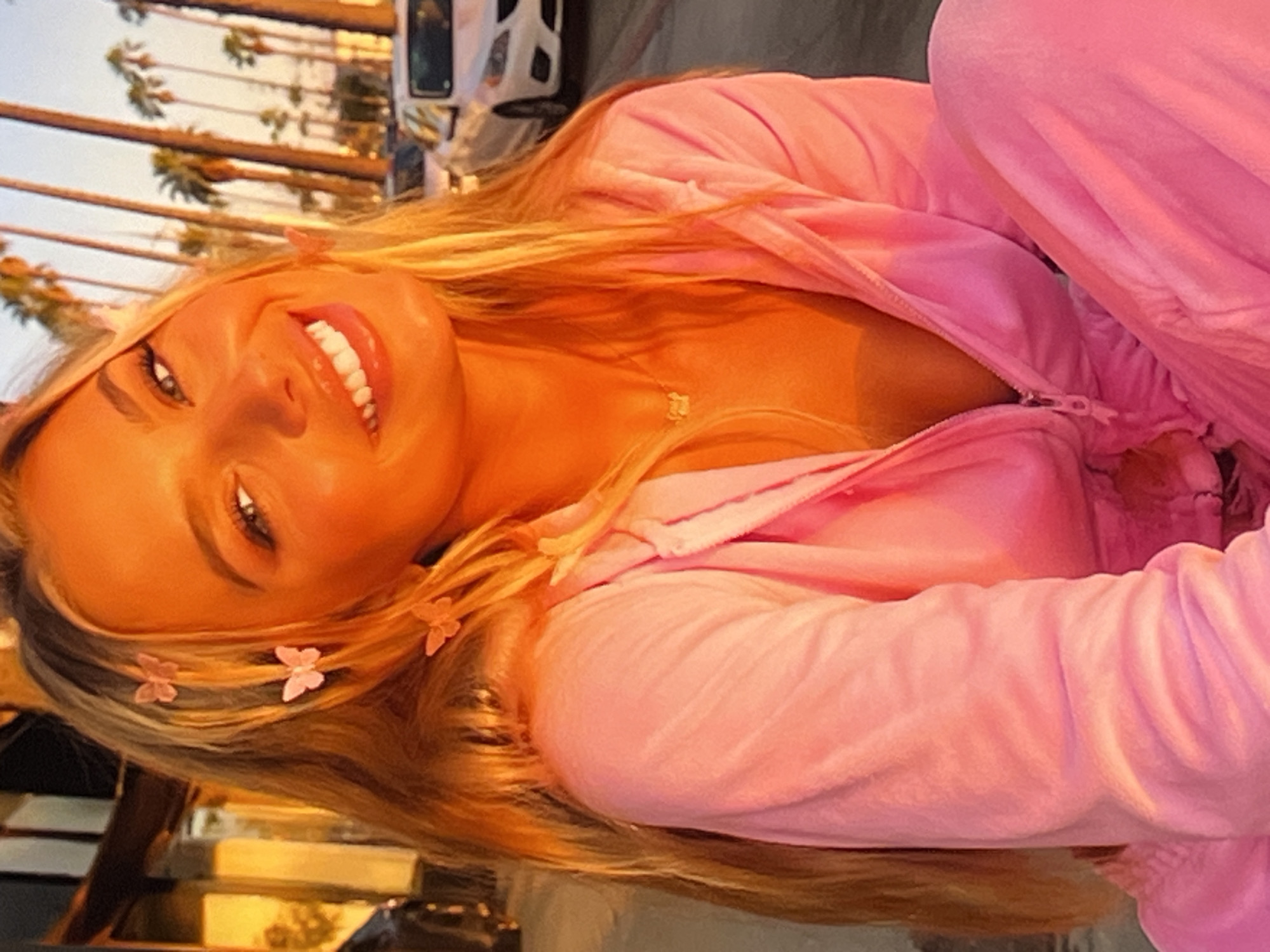 A young woman with blonde hair, adorned with pink butterfly clips, smiles broadly as she looks towards the viewer. She is wearing a bright pink velour tracksuit, with the zipper partially open. A delicate gold necklace with a small pendant is visible around her neck. The lighting suggests it is either sunrise or sunset, casting a warm, orangey glow on her face and the surrounding environment. In the background, a row of palm trees lines a street, with several white cars parked along the side. The scene appears to be in an urban setting, possibly a boulevard or a street known for its palm trees.