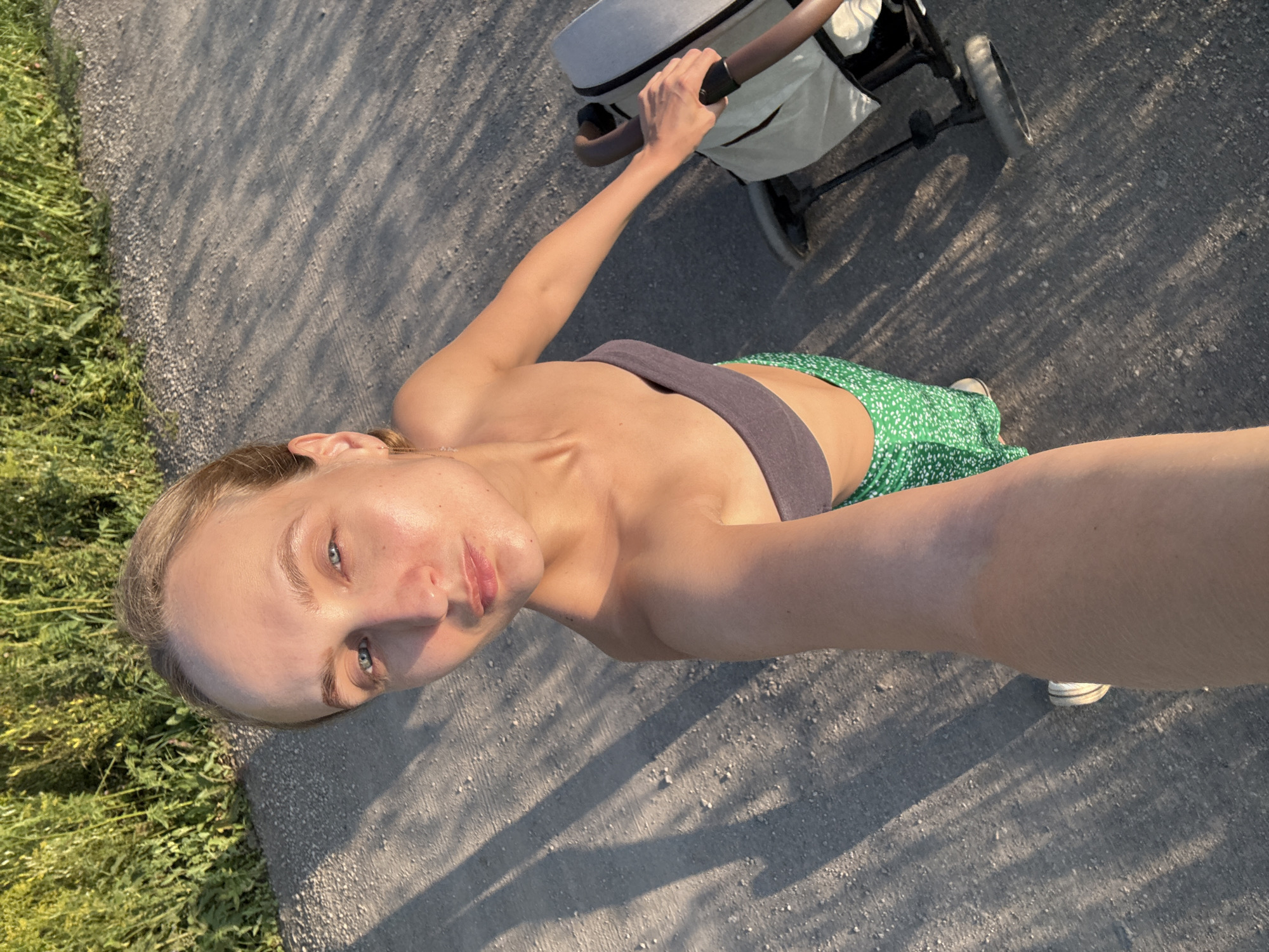 A woman is taking a selfie as she walks outdoors with a stroller. She is wearing a grey sports bra and a green and white floral skirt. Her blonde hair is pulled back, and she is looking at the camera. The stroller is a light grey color with a dark handle. The ground is a gravel path, and there is green grass on the left side of the image. Sunlight casts long shadows across the path, indicating it is likely late afternoon or early morning. The setting appears to be a park or a trail. The woman's expression is neutral. No text is visible in the image.