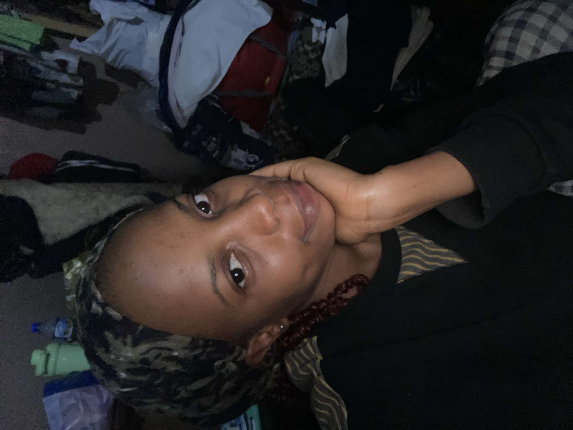 The image is a selfie of a young Black woman with dark skin and brown eyes, wearing a headscarf and a black long-sleeved shirt. She has a pensive expression and her hand is resting on her chin. The background is cluttered with clothing and other items, suggesting an indoor environment. The lighting is dim, creating a somewhat intimate and moody atmosphere. No specific landmarks or indicators of the city of Jos, Nigeria are visible in the image.
