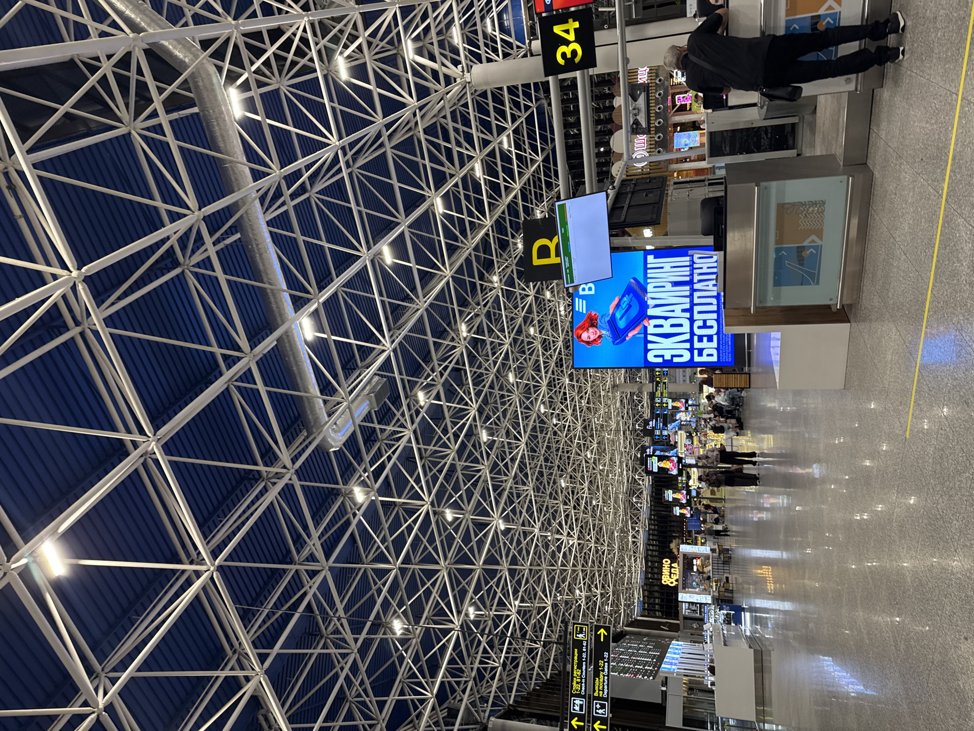 This is a photograph of the interior of Sochi International Airport, located in Sochi, Russia. The focal point of the image is the airport's ceiling, which features a complex network of white metal beams forming a geometric, grid-like pattern. Blue panels are visible behind the beam structure.
The image includes signage with Cyrillic text, and the number 34. There are departure gates, check-in counters, and information displays. People are present in the scene, and at least one person is interacting with a counter or kiosk. The floor is shiny, reflective, and appears to be made of polished stone.