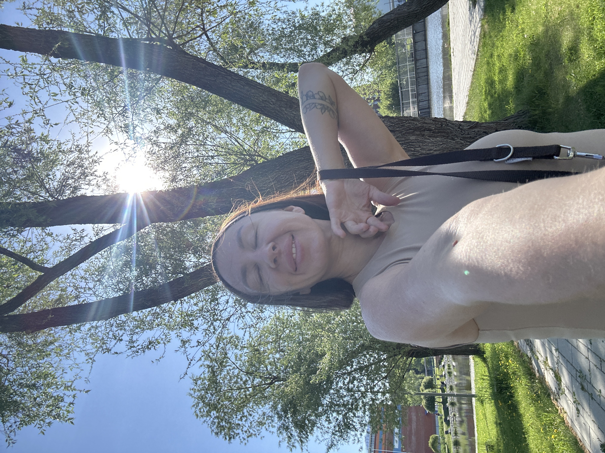 A woman takes a selfie under trees in Votkinsk, Russia, on a sunny day. The sun is directly overhead. She is wearing a beige top and has a tattoo on her left arm. She is holding a black leash in her hands. A canal is visible in the background. The ground is covered with grass and a paved walkway is next to the grass.