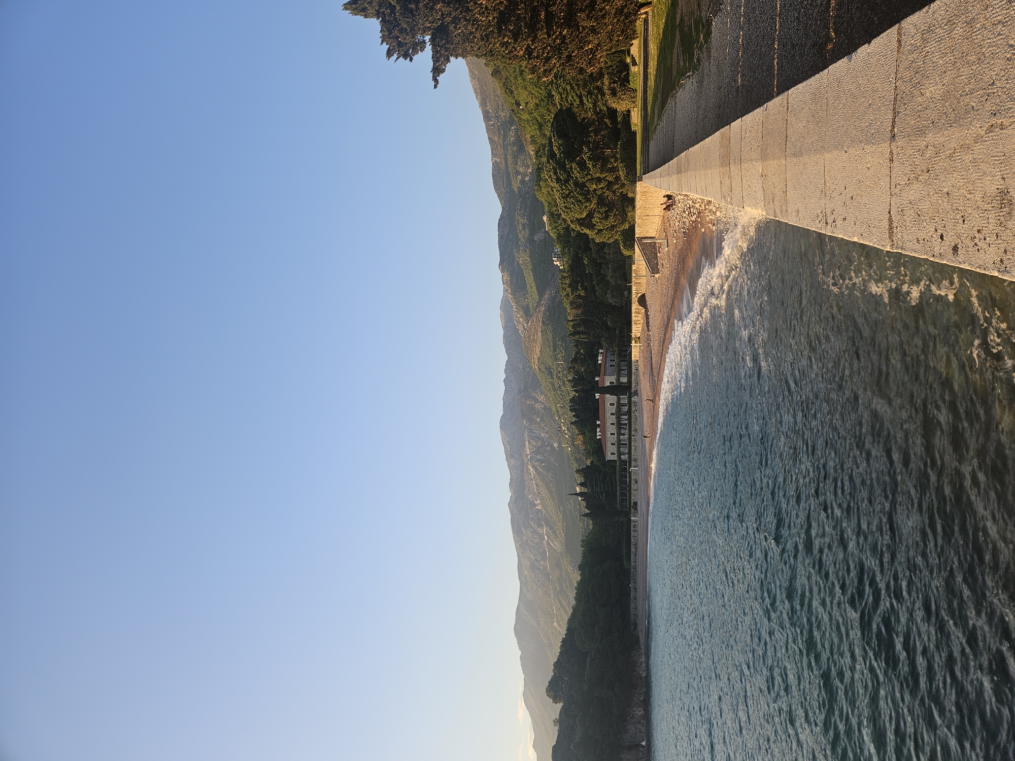 This wide landscape shot captures a serene coastal scene under a clear blue sky, likely during late afternoon or early morning due to the warm, golden light. In the foreground on the right, a long, light-colored concrete or stone pier/wall extends into the distance, brightly illuminated by the sun. Gentle waves with white foam break along its edge and lap onto a narrow, pebbly beach immediately adjacent to it.
The sea, a deep blue-green, occupies the lower right portion of the frame, exhibiting ripples and textures on its surface. Moving towards the left and center, the coastline features lush, dark green vegetation, including numerous trees and bushes, covering hills that gradually rise into a range of majestic mountains. These mountains dominate the background, appearing somewhat hazy in the soft light.
Nestled among the trees along the coast, several multi-story buildings with light-colored facades and reddish roofs are visible, suggesting a resort or residential area. Small, indistinct figures can be seen on the beach and possibly on the pier, indicating human presence. The sky, a vast expanse of light blue, occupies the upper left section of the image, devoid of clouds. The overall impression is one of peaceful natural beauty and tranquility. There is no visible text in the image.