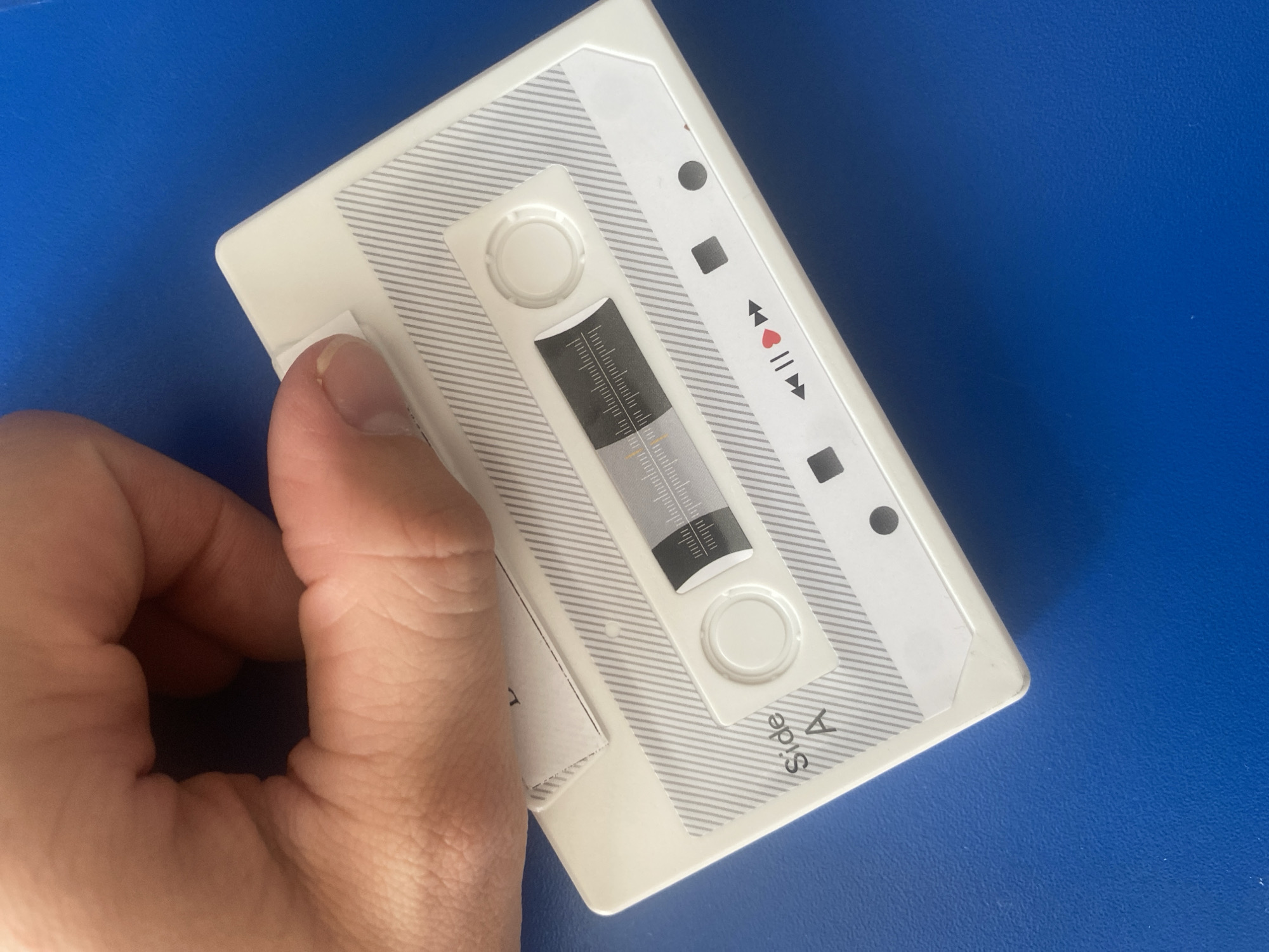 A person's hand is holding a white cassette tape with a gray striped pattern. The tape has a clear window showing a black and white scale and two reels. There are also symbols for play, pause, and rewind on the tape. The text "Side A" is visible on the left side of the tape. The background is a bright blue surface. There is no information to determine the location in Vitsebsk, Belarus.