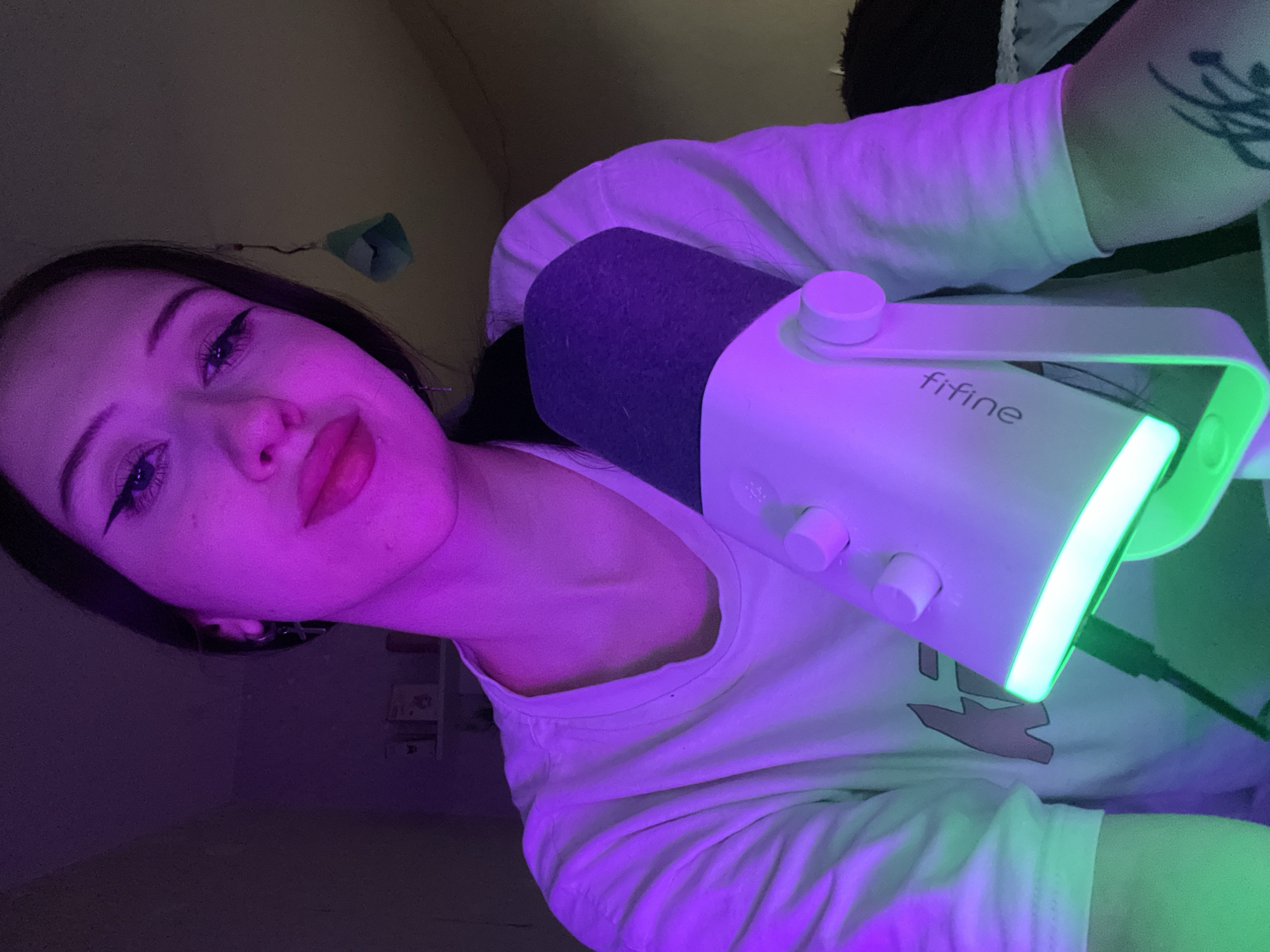 The image shows a close-up of a young woman with dark hair and dramatic winged eyeliner, wearing a white t-shirt. She is positioned in front of a white and purple microphone with a green LED light strip. The lighting in the scene is primarily purple and green. The background is dark and indistinct, with some shelves and objects visible on the left. The woman's lips are painted a vibrant pink. There is a tattoo visible on her arm. The image does not contain any identifiable landmarks or clues that would suggest a specific location within Votkinsk, Russia.