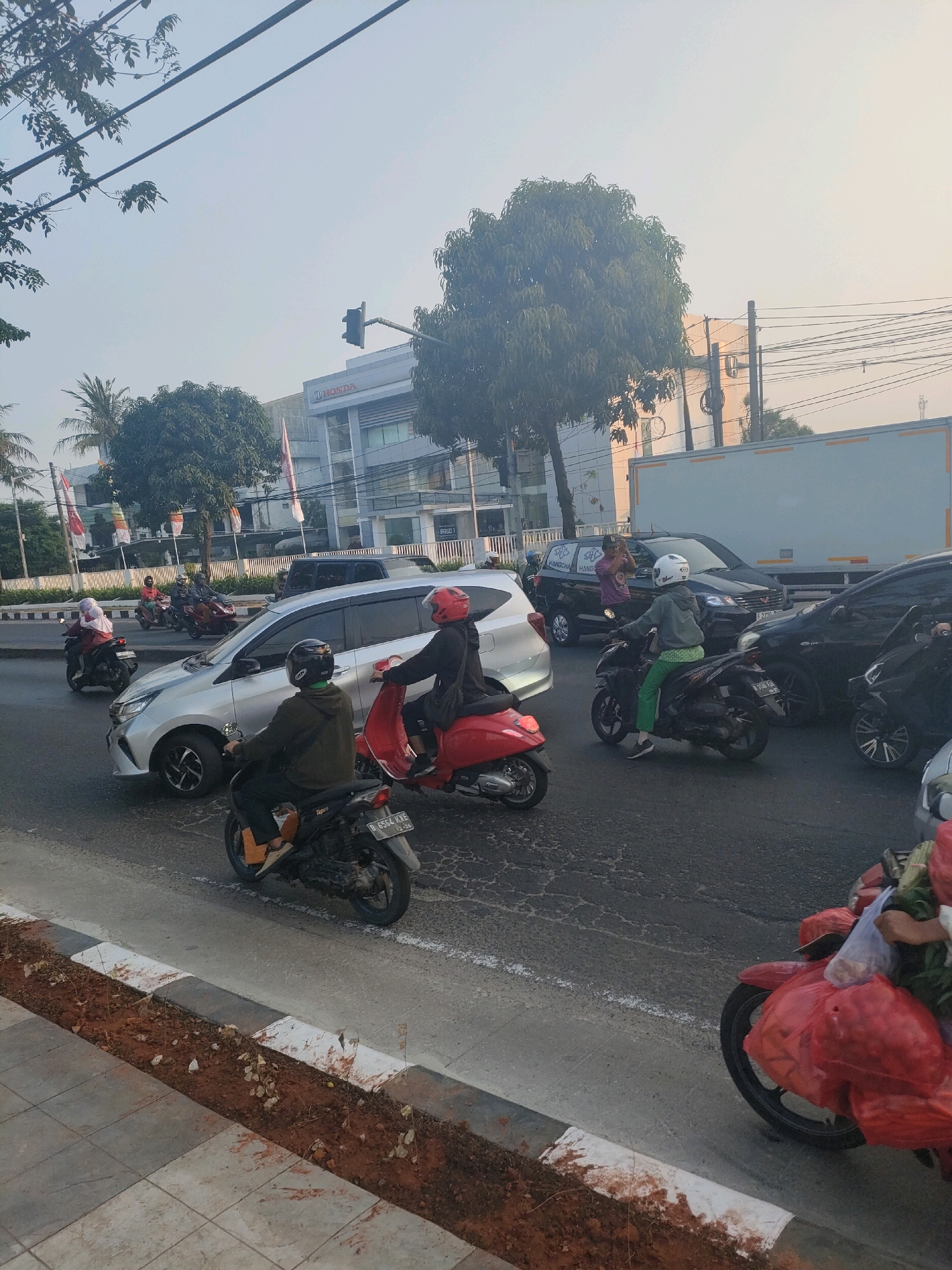 This is a daytime, medium-shot, eye-level photograph taken on a street in Bekasi, Indonesia. The scene depicts traffic congestion with several motorbikes and cars on a multi-lane road. The street has a clear white line marking the lanes and a curb with a dirt median. In the background, a multi-story building with a "HONDA" sign is visible, along with trees and power lines. Several people are riding motorbikes, some wearing helmets and casual clothing. One motorbike has a license plate that reads "B 6564 KXE". Another person is on a red scooter, and a person in the foreground is on a motorbike carrying red bags. The sky is hazy, suggesting a warm, possibly humid day. The overall impression is of a typical urban traffic scene.