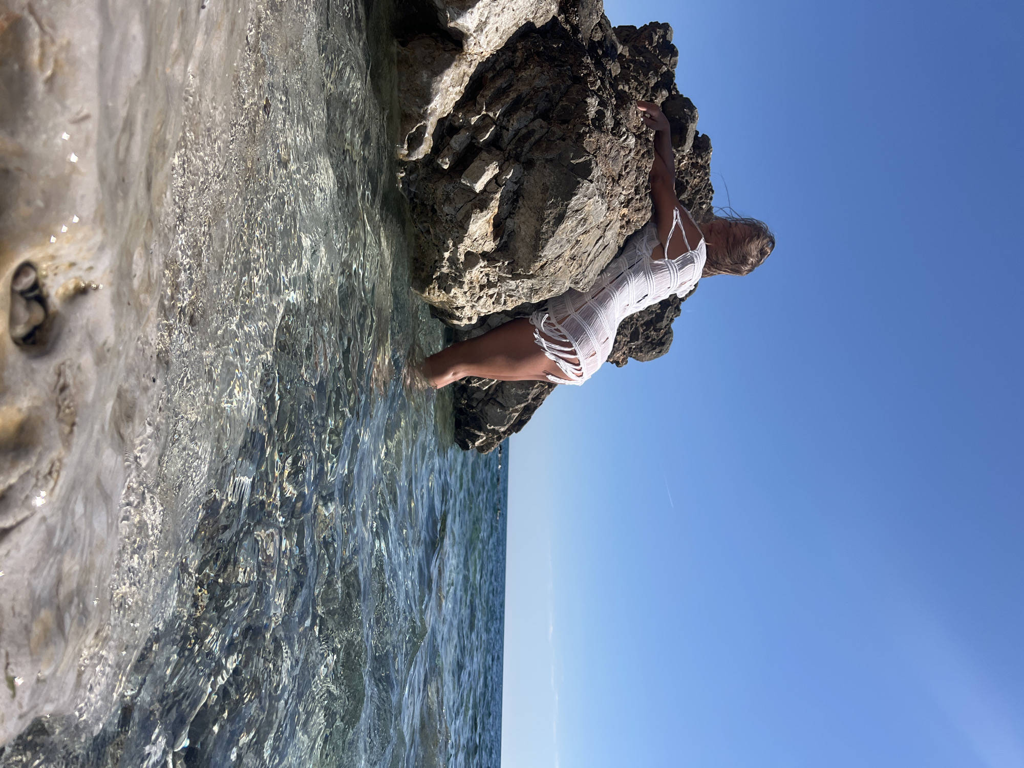 A woman wearing a white, fringed dress is leaning against a rocky outcrop at the edge of clear, shallow water. Part of her legs and feet are submerged. The outcrop is dark and rough in texture. The water is clear and reflects the sky. The sky is a bright, cloudless blue. The location is Prijevor, Montenegro.