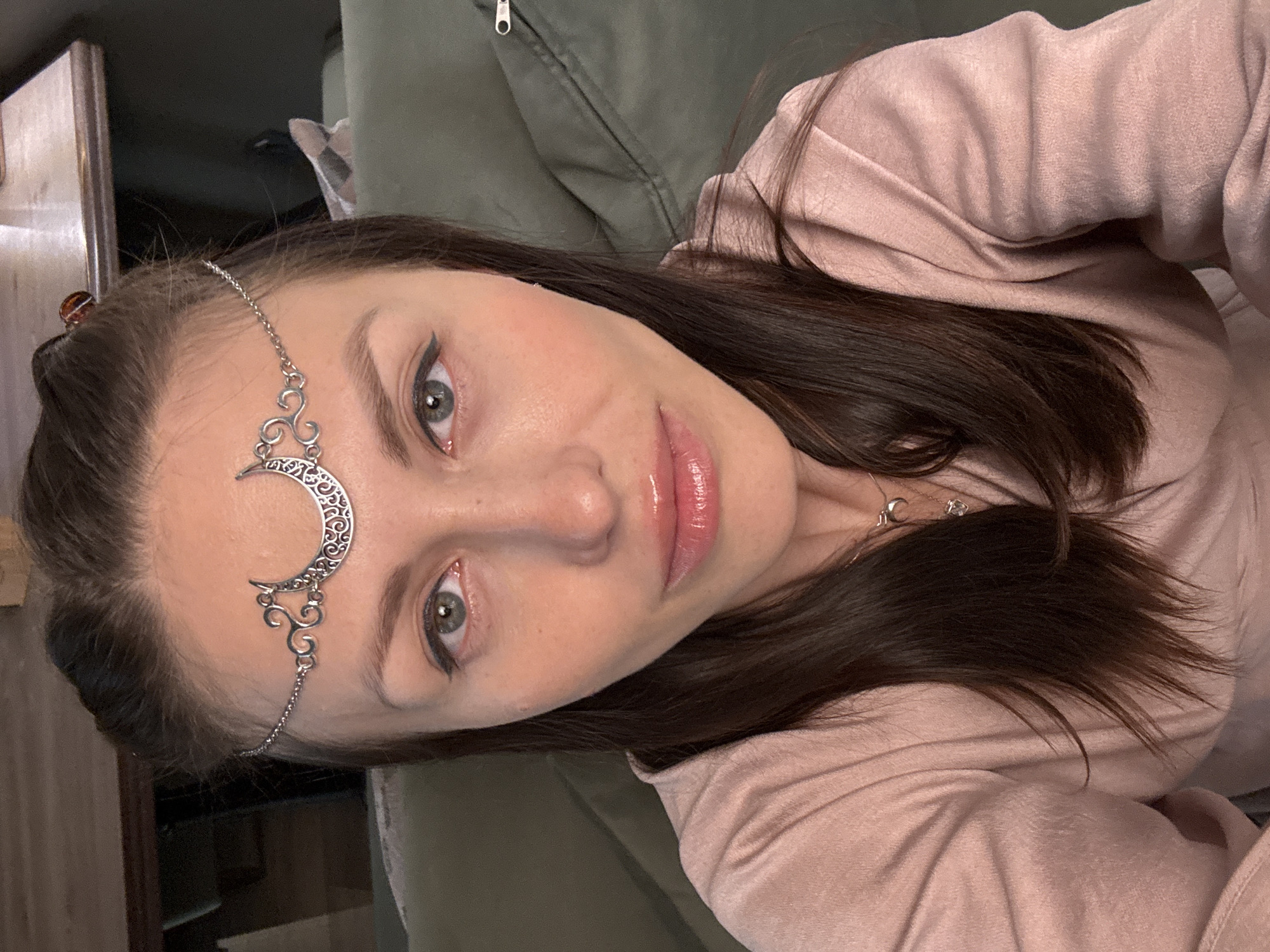 A woman with fair skin, green eyes, and dark brown hair poses for a selfie. She is wearing a silver crescent moon headpiece. Her makeup includes eyeliner and glossy pink lipstick. She is wearing a pink top and a silver necklace with charms. The background is a green upholstered surface and a wooden cabinet. The location is Unknown, Russia.
