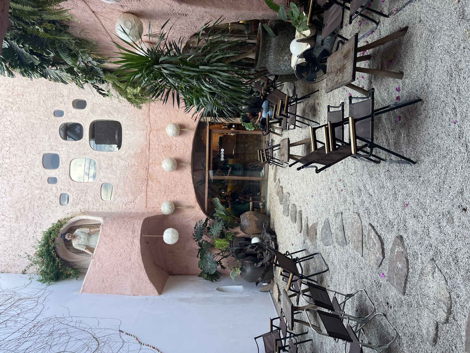 The image shows an outdoor seating area in an unknown city, with the camera positioned to view the space in a vertical format. The architecture has a unique, slightly surreal design featuring pinkish walls with various rounded windows and an arched entrance. On the left side of the frame, a woman is visible in a circular alcove, adorned with greenery. The seating area is set on light-colored gravel, with stepping stones creating a pathway. Several chairs and small tables are arranged for patrons. Lush greenery, including tropical plants, is interspersed among the tables. People are seated in the distance, suggesting a relaxed, social setting. Lanterns are attached to the walls.