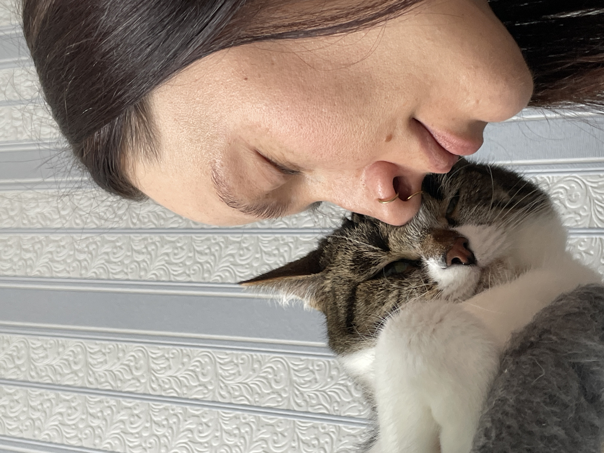 A woman with dark hair, a nose ring, and closed eyes is positioned next to a tabby and white cat. The woman's face is close to the cat. The cat's eyes are also closed, and it appears to be resting or being held. The background is a textured, patterned surface, which seems to be a wall. There is no evidence to suggest this photo was taken in Izhevsk, Russia.