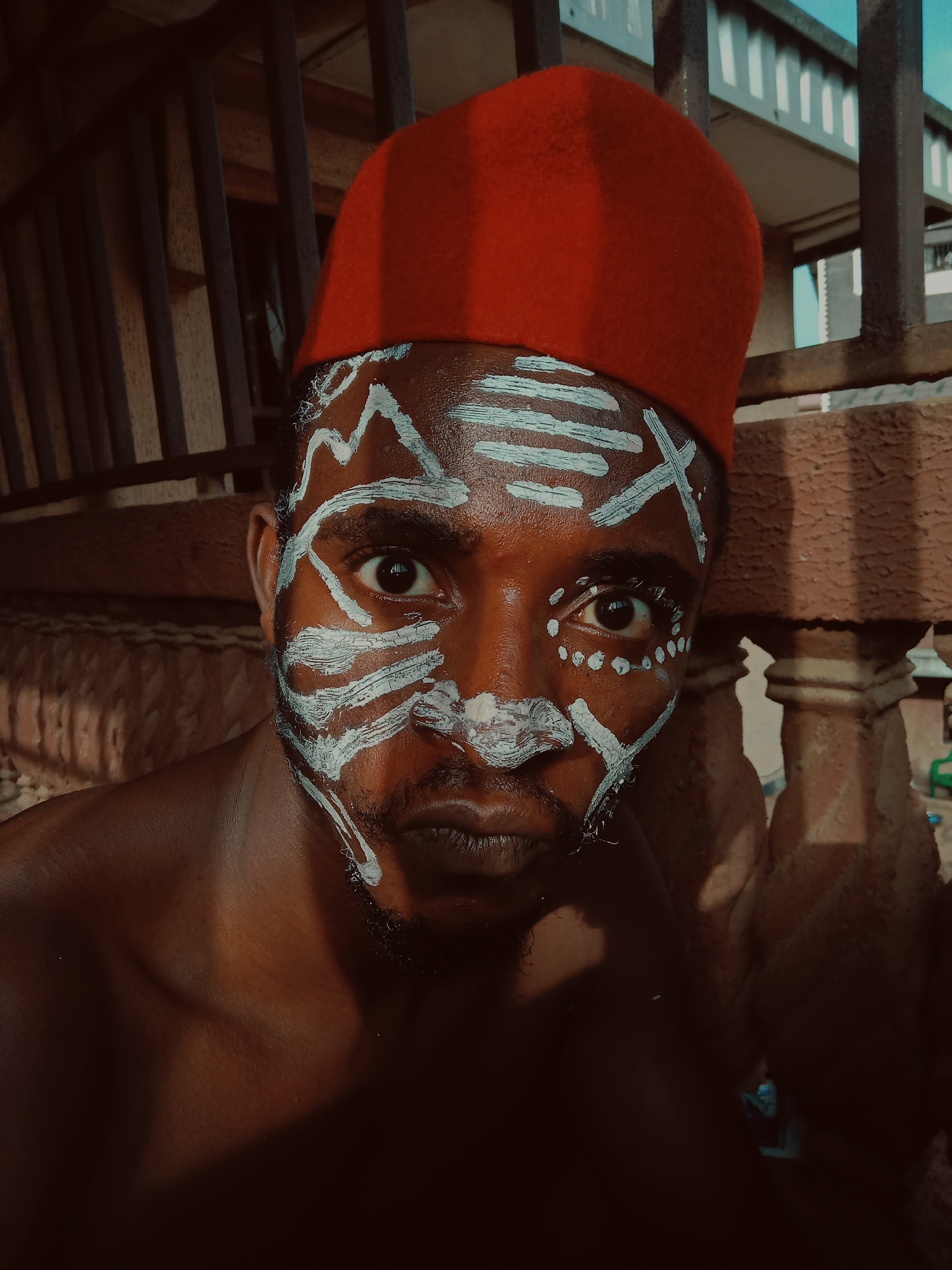 A close-up shot captures a dark-skinned man in Nkpor, Nigeria. He wears a bright red fez hat and has white tribal-like markings painted on his face. His face is expressive, his dark eyes looking directly at the viewer. He stands in front of a metal fence with a concrete pillar. He appears shirtless.