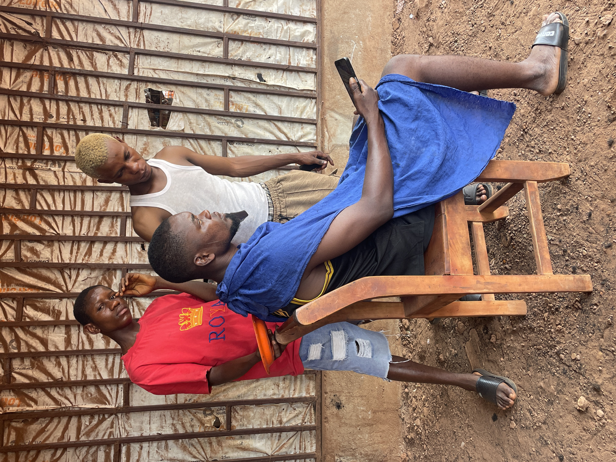 Three young Black men are gathered outdoors in a casual setting, likely in Ogbe, Nigeria. The ground is covered with reddish-brown earth and small stones. Behind them stands a makeshift wall constructed from light-colored sheeting or packaging material, possibly plastic or fabric, framed by wooden beams. Text visible on the wall includes "Muntajat" and "25KG".
Two of the men are seated on simple wooden chairs. The man on the right is partially reclined, wearing black shorts and a blue cloth draped over his lap. He is holding a black smartphone with both hands, engrossed in its screen. He wears black slides, one of which clearly displays "CONVE" (likely "CONVERSE").
Beside him, a man with short, bleached blonde hair stands wearing a white tank top and brown patterned shorts. He looks downwards with a neutral expression.
On the left, the third man is seated, wearing a red t-shirt featuring the word "ROYAL" and a crown graphic. He has ripped light blue denim shorts and black slides, holding an orange plate or frisbee in his hands. He looks towards the viewer with a calm expression.
The scene suggests a relaxed daytime gathering in an informal outdoor environment.