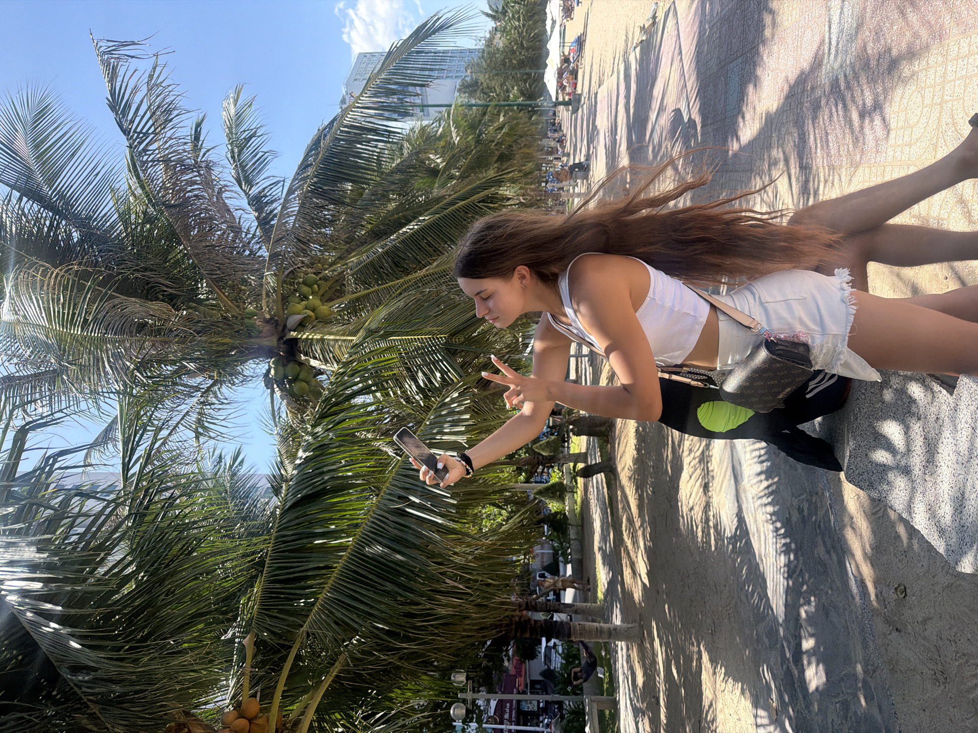 In Nha Trang, Vietnam, a young woman with long, brown hair is captured taking a selfie outdoors, near a beach. She is wearing a white tank top and denim shorts. She is holding a smartphone in her right hand, and her left hand is extended, forming a peace sign. A dark-colored crossbody bag with a patterned design is slung over her shoulder. She is positioned in front of several palm trees, some of which bear coconuts. Behind her, a paved walkway lines the beach, with lounge chairs and umbrellas visible in the distance. Buildings, including a tall, modern structure, are also present in the background. The ground is a mix of sand and patterned paving stones, with palm fronds casting shadows. A person is visible lying in a hammock strung between two trees in the mid-ground, and other figures are seated on benches further down the walkway. The sky is clear and blue.