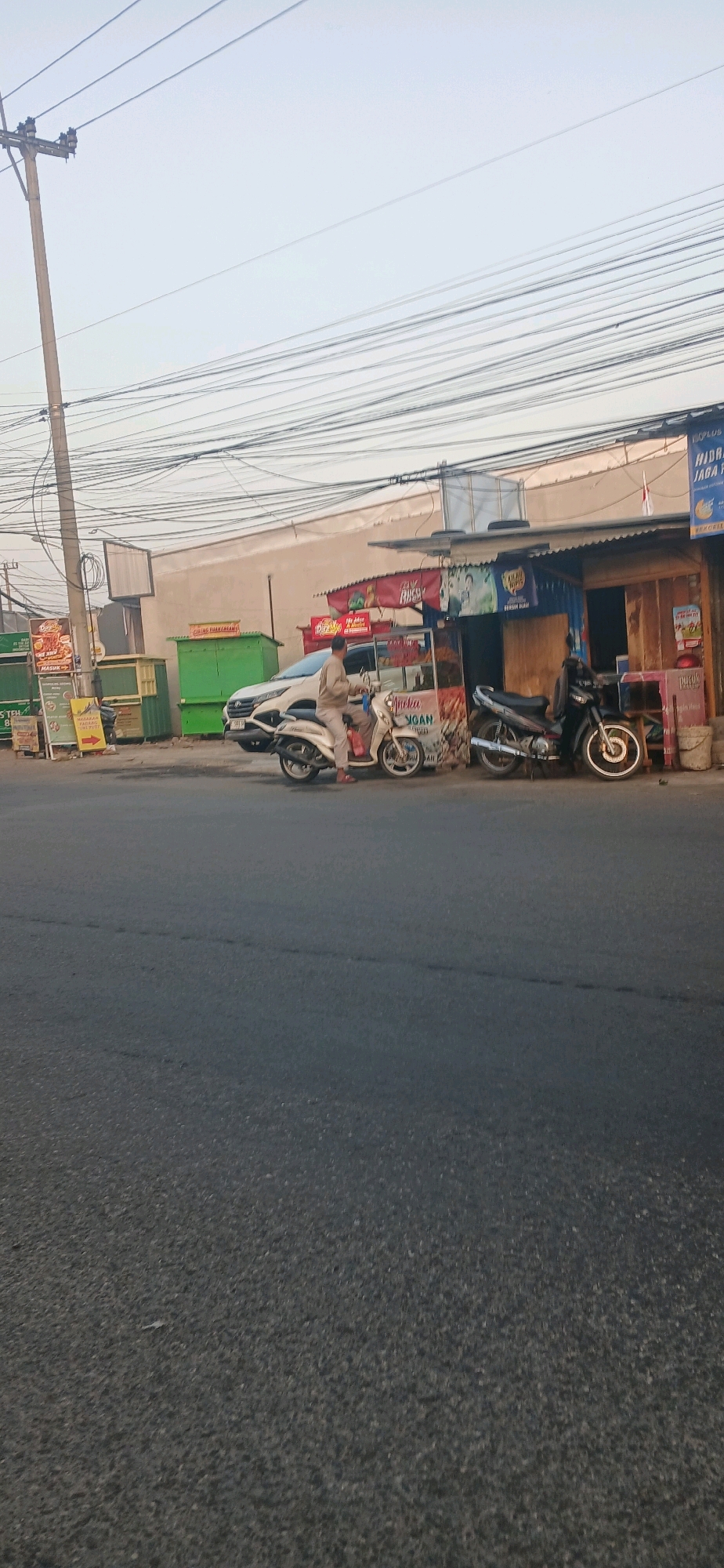 A street scene in Cirebon, Indonesia, features a vendor cart on the left with a person standing next to it. Two motorcycles are parked on the right. A white car is parked behind the vendor cart. The scene is bordered by a wall on the right and a utility pole with numerous wires on the left. The sky is overcast.