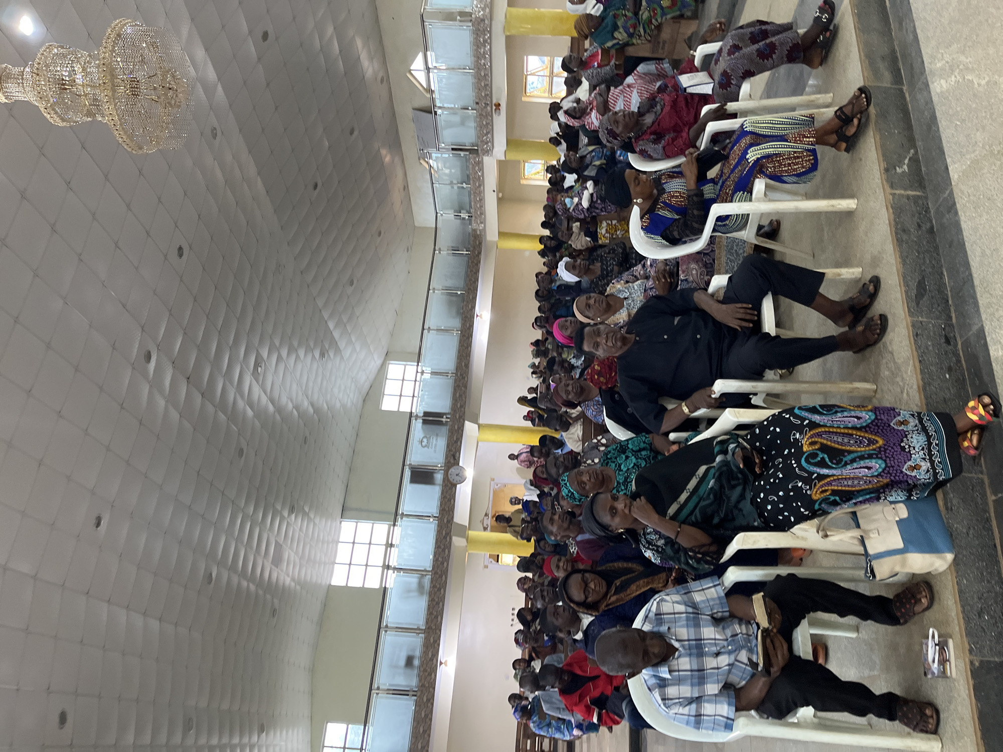 The image shows an indoor gathering of people seated in rows of white plastic chairs. The location is identified as Jos, Nigeria. The attendees are dressed in a variety of clothing styles, including traditional African prints. The environment appears to be a large hall with a high ceiling, featuring a decorative chandelier and windows along one side. People are seated attentively.