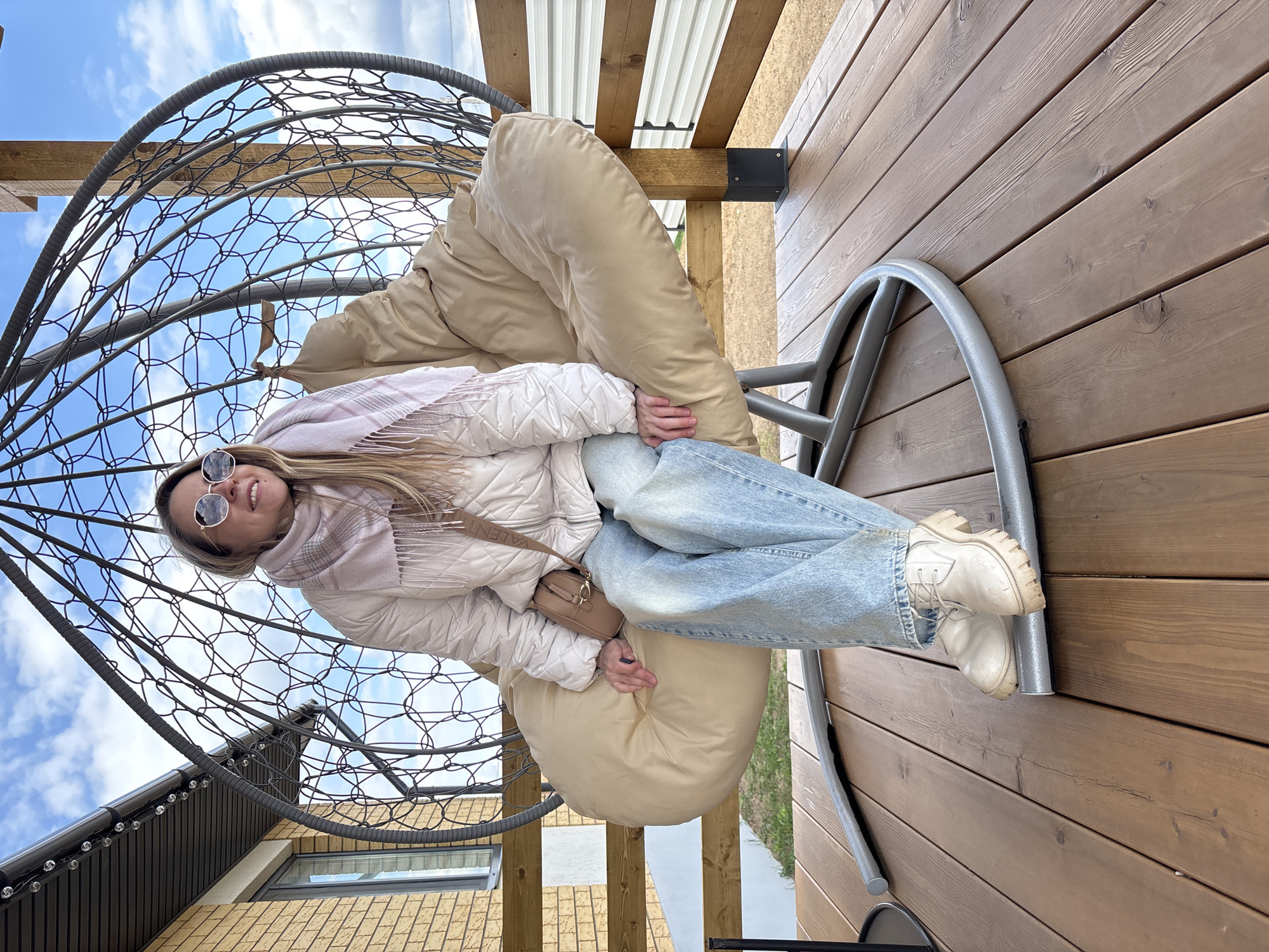 A young adult woman with long blonde hair, wearing round sunglasses, a white quilted jacket, a light pink patterned scarf, light blue jeans, and white chunky-soled boots, is shown smiling brightly at the camera. She is seated comfortably in a large, dark metal hanging egg chair with a plush beige cushion. Her hands are resting on her lap, and she appears to be holding a small dark object. A small brown shoulder bag, with the text "VALENTINO" visible on its strap, is worn across her body.
The setting is outdoors on a wooden deck or balcony. The flooring is composed of warm brown wooden planks. In the background, a body of water and distant greenery can be seen, suggesting a scenic overlook. Above her, a structure with corrugated metal roofing and wooden beams is visible. A building featuring light brown brickwork and dark siding is partially visible to her left. The sky is blue with scattered white clouds, indicating a bright daytime. The woman's relaxed pose and cheerful expression convey a sense of enjoyment. The image is presented rotated 90 degrees clockwise from its natural orientation.