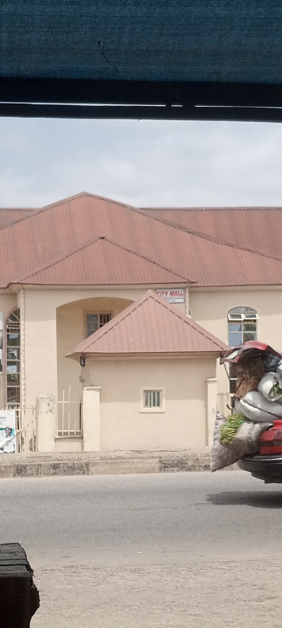 The image shows a building identified as "CITY MALL" in Jalingo, Nigeria. The building has a multi-level, reddish-brown metal roof and a beige facade. A smaller, attached structure also has a reddish-brown metal roof. There are several windows visible on the building, including arched windows on the upper level and smaller rectangular windows. A white metal fence is present in front of a section of the building. On the right side of the image, a vehicle's trunk is loaded with sacks and bundles of produce, suggesting a market or transportation activity. The foreground shows a paved road and a curb. The overall scene depicts an urban commercial area with a mix of building architecture and street-level activity.