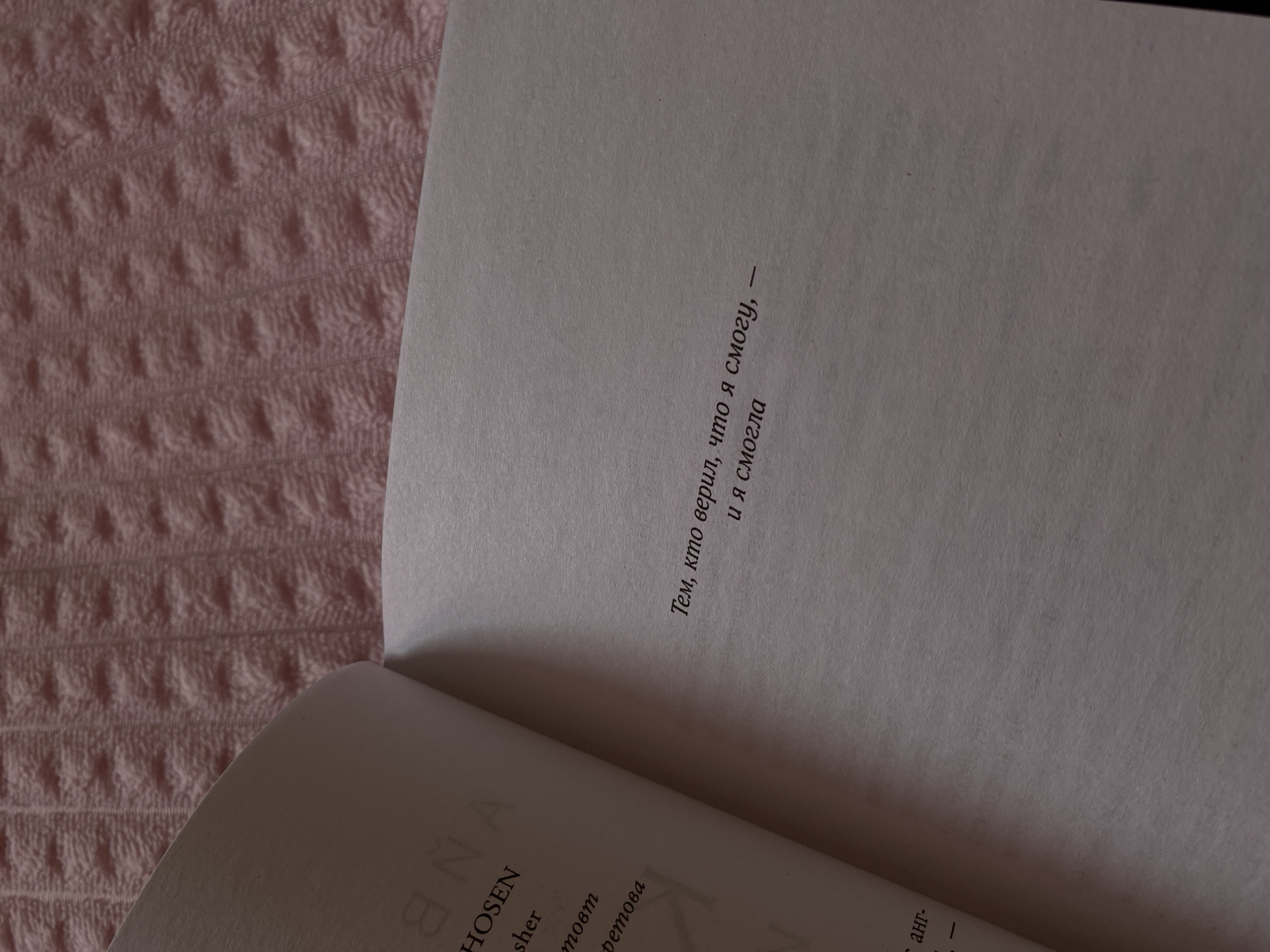 The image shows an open book lying on a pink, textured surface, possibly a blanket or bedspread. The book's right-hand page contains a short text written in Russian: "Тем, кто верил, что я смогу, и я смогла", which translates to "To those who believed I could, and I did." The left-hand page is partially visible and displays some text in a Latin alphabet, with the words "NA HOSEN sher товт Детова" partially readable. The lighting is soft and casts shadows across the page, emphasizing the texture of the paper and the surrounding surface. The scene is indoors, likely in a private residence or cozy setting, but it's not possible to pinpoint the specific location in Votkinsk, Russia, without additional information.