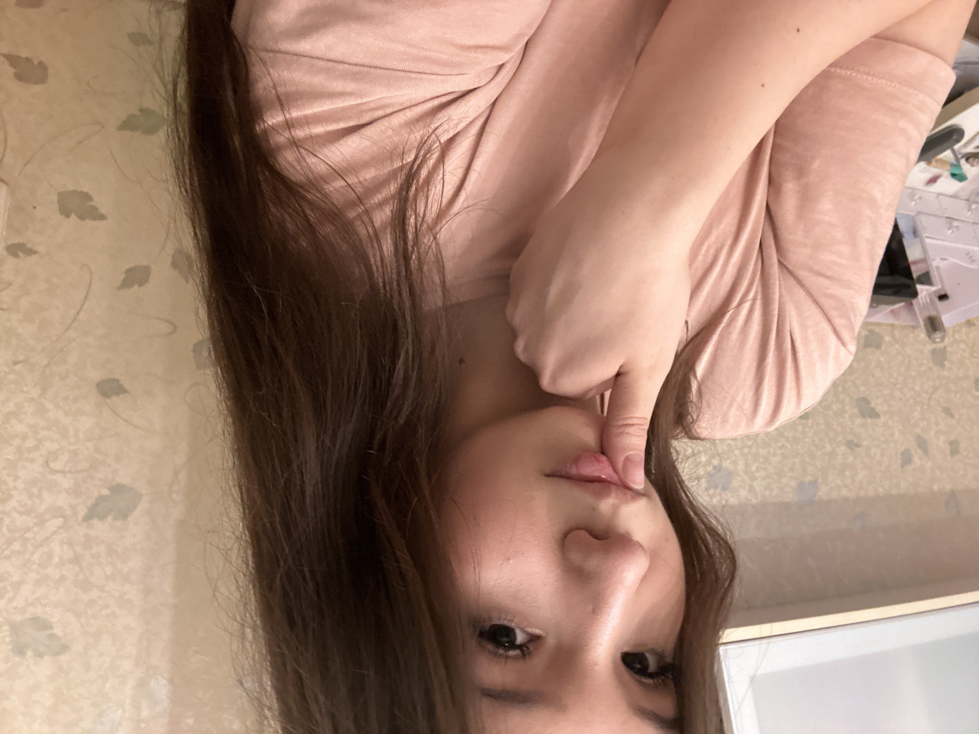 A young woman with long, dark brown hair is depicted in a close-up indoor portrait. She is wearing a light pink or peach-colored long-sleeved shirt. Her head is slightly tilted, and she looks directly at the camera with a direct and thoughtful gaze. Her right index finger is placed against her lower lip. The background features light-colored wallpaper with a subtle, recurring pattern of abstract leaf shapes on the left side of the frame. The right side of the background is blurred, showing light surfaces that suggest an indoor setting, possibly a desk or shelf with indistinct objects. There is no visible text.