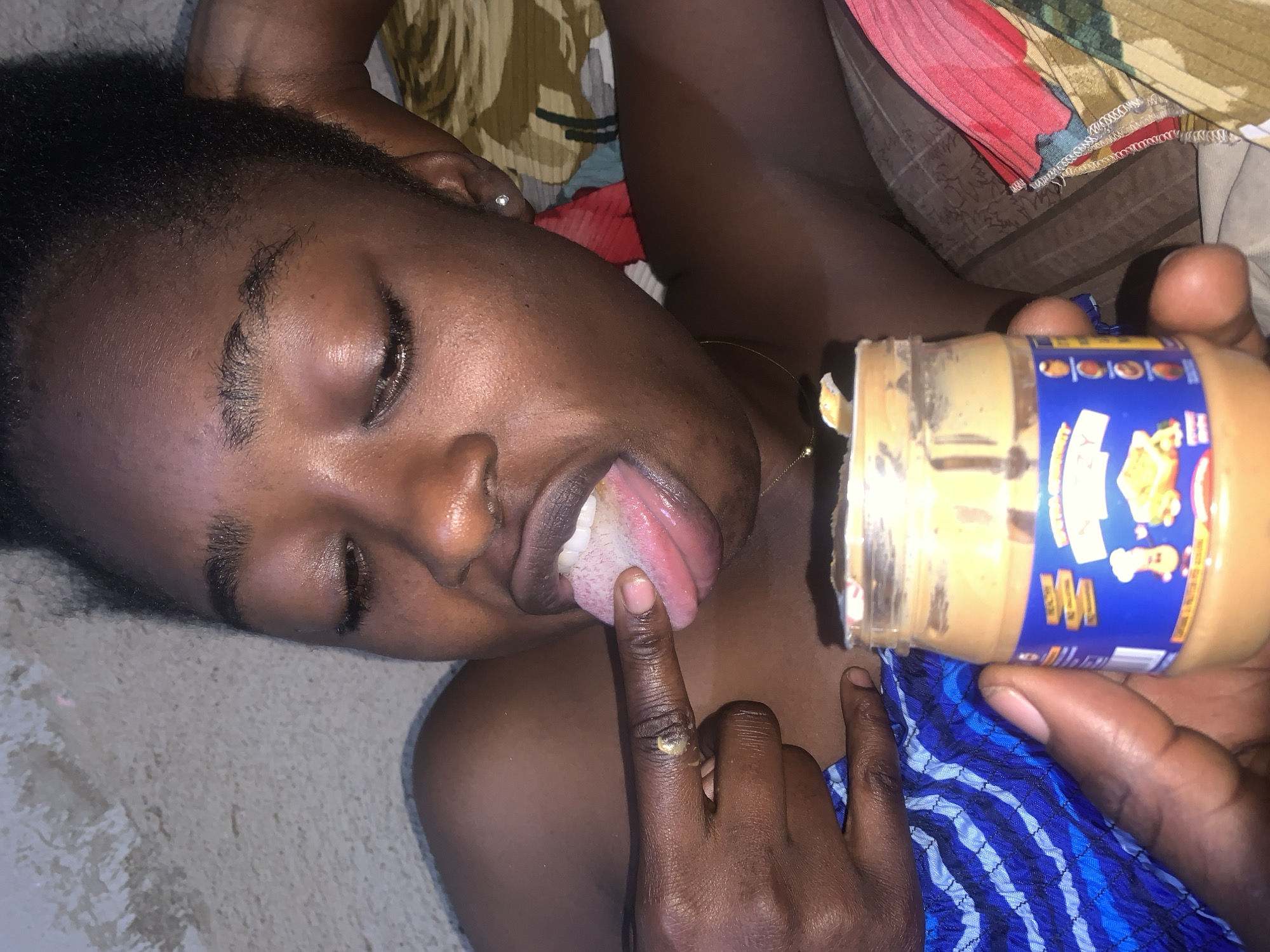 The media file shows a close-up of a person lying down, with their eyes closed and tongue sticking out, in what appears to be a home setting in Rafin Par, Nigeria. The person's face is the primary focus, with their mouth open and tongue extended towards a jar of peanut butter. The person's right index finger, which has some peanut butter on it, is also touching their tongue.
Objects visible include the jar of peanut butter, which has a blue label with some text and images on it, and a ring on the person's right index finger. The person is wearing a necklace and what appears to be a blue and white patterned fabric. The background is blurred, showing what could be bedding or clothing with floral patterns.
The activity depicted is the person interacting with the peanut butter, seemingly tasting it directly from the jar or their finger. The lighting suggests it could be daytime, but there are no specific clues about the time of day or weather. The overall mood is playful or experimental, given the action.
Visible text on the peanut butter jar is in blue and features some cartoon images and text. It is difficult to read the specific brand or details of the text due to the resolution and angle. The location context provided is Rafin Par, Nigeria.
