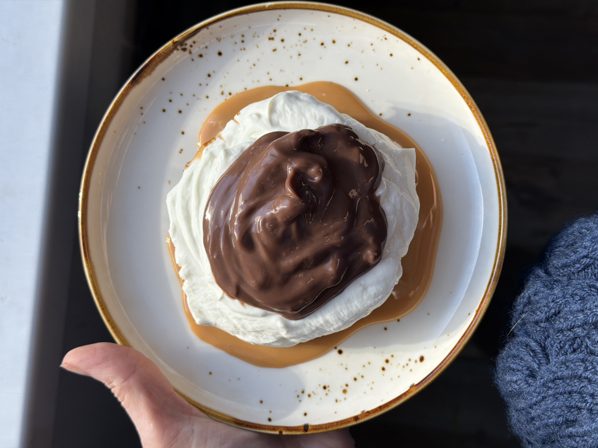 A top-down shot shows a plate of dessert held by a hand wearing a blue knitted sleeve. The dessert consists of a light brown liquid (possibly caramel) spread on the plate, topped with a dollop of white whipped cream, and then covered with a shiny, dark brown chocolate topping. The plate is white with a brown rim and scattered brown speckles. The background is blurred. The location is not possible to identify.