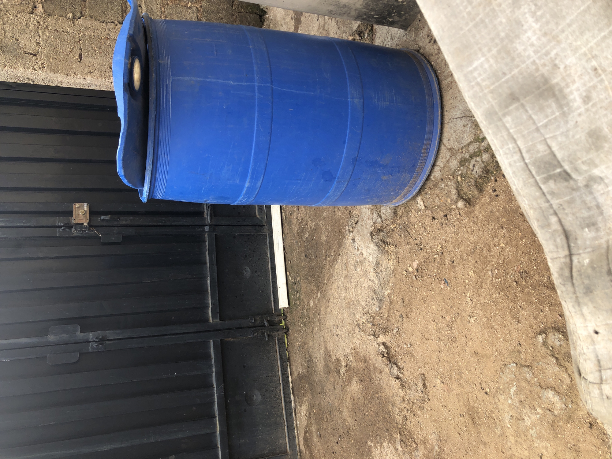 The scene captures an outdoor area in Kabong, Nigeria, featuring a dark metal gate, a blue plastic barrel, and an unpaved ground surface.
The central and left portion of the image is dominated by a dark, possibly black or dark grey, corrugated metal double gate. The gate panels are secured by a central latch mechanism. On the right panel of the gate, an external locking device is visible, comprising a padlock attached via a chain, positioned adjacent to a crude electrical box or switch with exposed wiring. A segment of white plastic piping or conduit is laid horizontally along the base of the gate on the ground.
To the right of the gate, a large, upright blue plastic barrel stands on the unpaved ground. The barrel features horizontal molded ridges along its circumference. Its lid is partially open, resting askew on the top rim, revealing the interior opening. Behind and to the right of the barrel, a light brown or grey rough-textured wall constructed of concrete blocks is visible.
The ground surface in the foreground and mid-ground consists primarily of uneven reddish-brown earth mixed with small stones and drier, lighter-colored patches. The bottom right corner of the image is partially obscured by a blurry, light-toned, textured object, possibly fabric or canvas, extending diagonally into the frame. No individuals or active interactions are present in the scene.