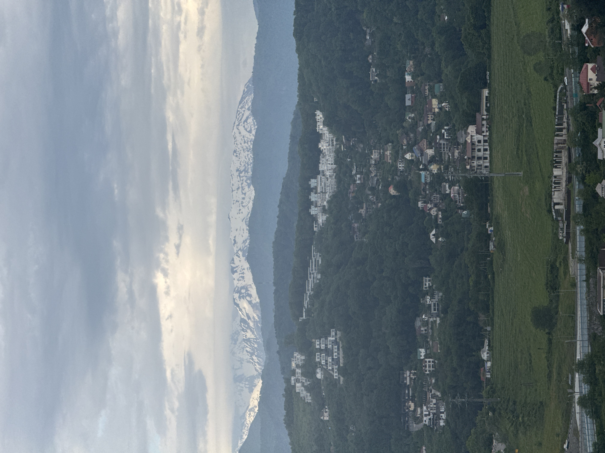 The image shows a mountainous landscape with a town nestled on the slopes. In the distance, a snow-capped mountain range is visible under a cloudy sky. Closer to the viewer, a densely forested hillside is dotted with buildings, suggesting a residential area. The terrain transitions to a flatter, green area with more houses and structures, possibly a residential or suburban setting. The image is rotated 90 degrees clockwise. There is no indication that the photo was taken in Sochi, Russia.